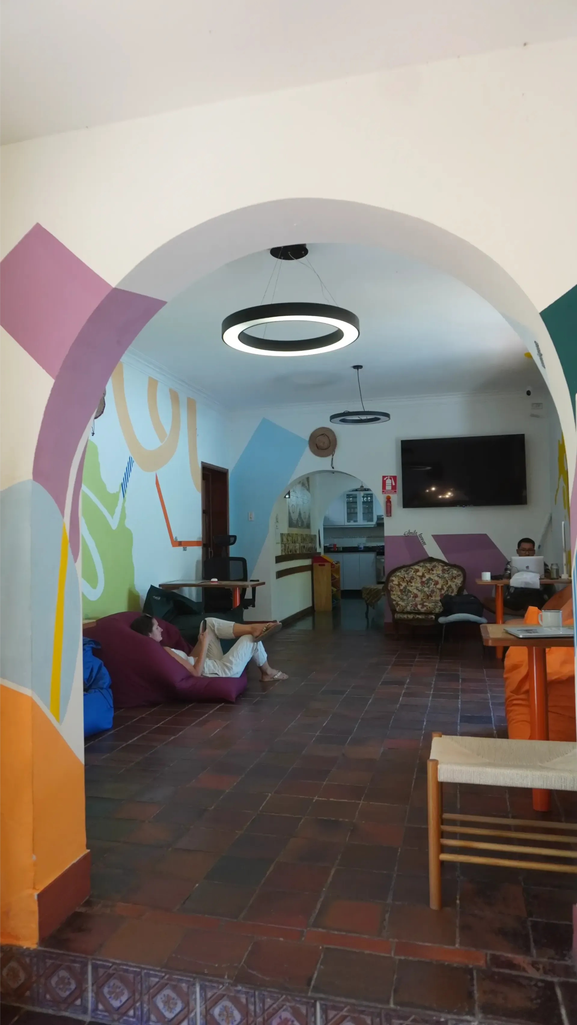 View through an archway into a colorful room. Two people relax on the floor, a TV, and furniture are visible.
