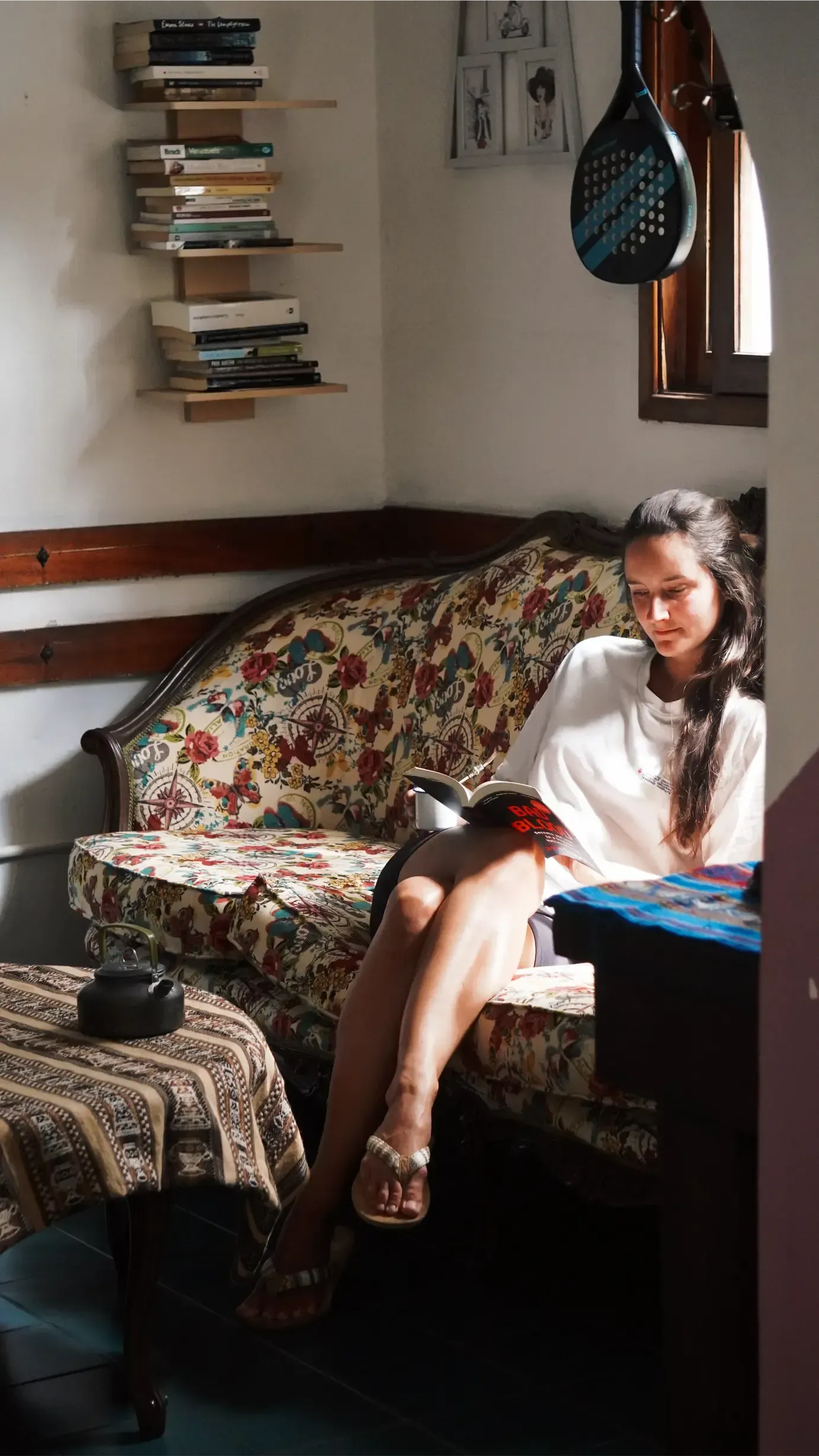 Woman reading on floral sofa in sunlit corner; books on shelves above.