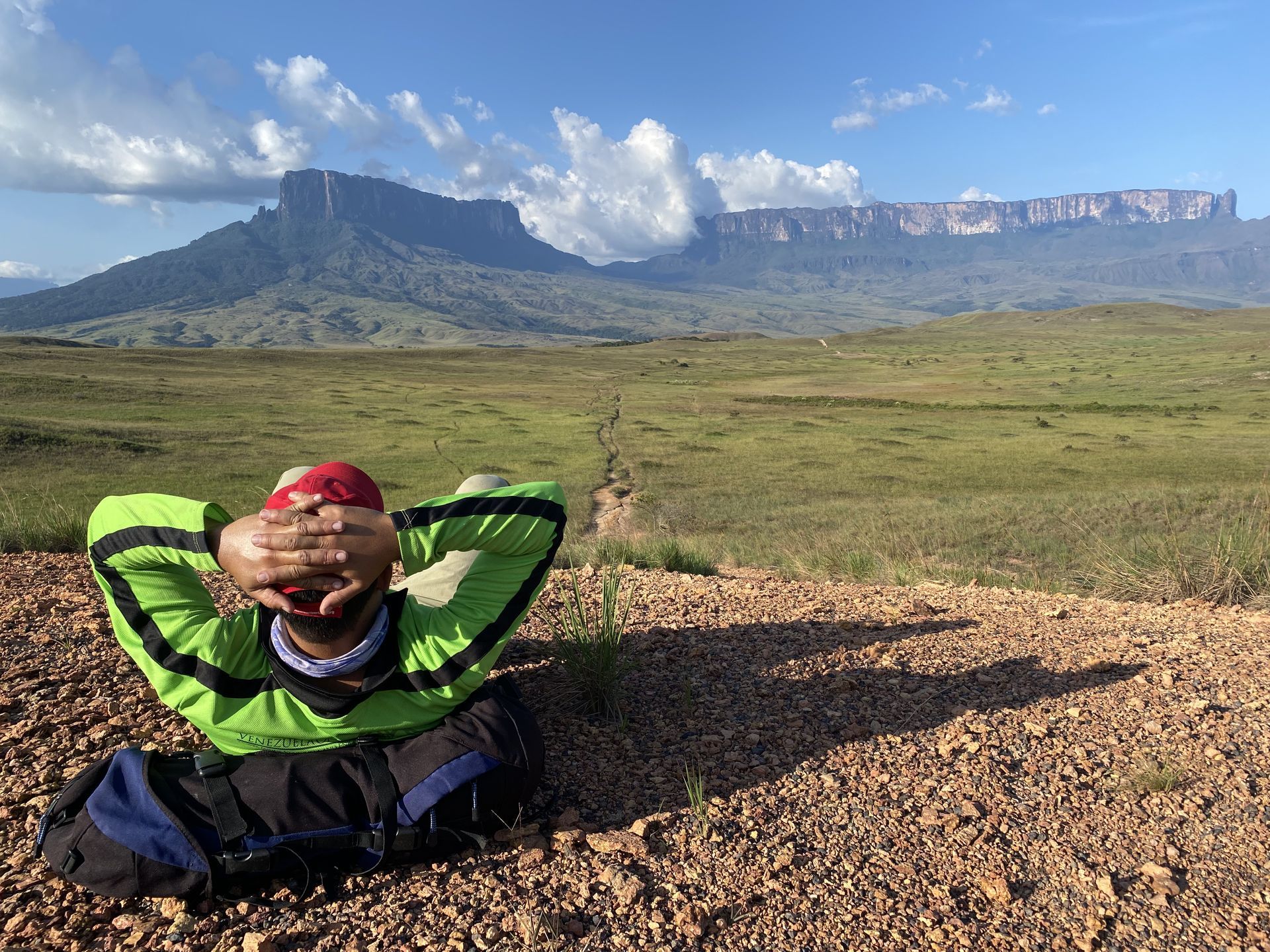 Person resting on dirt path, arms behind head, with large flat-topped mountains and grassy plain in background under a blue sky.