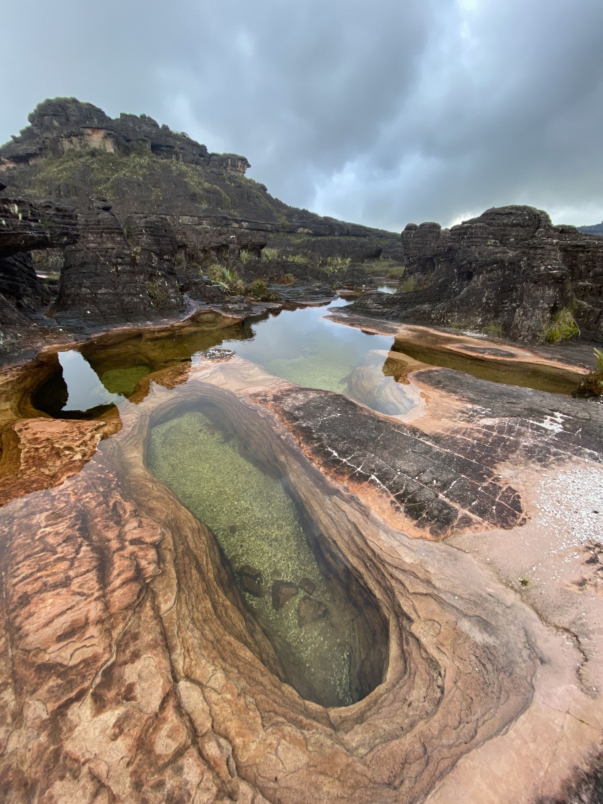Rocky landscape with several pools of water reflecting the cloudy sky. The reddish-brown and dark rocks contrast with the clear water.