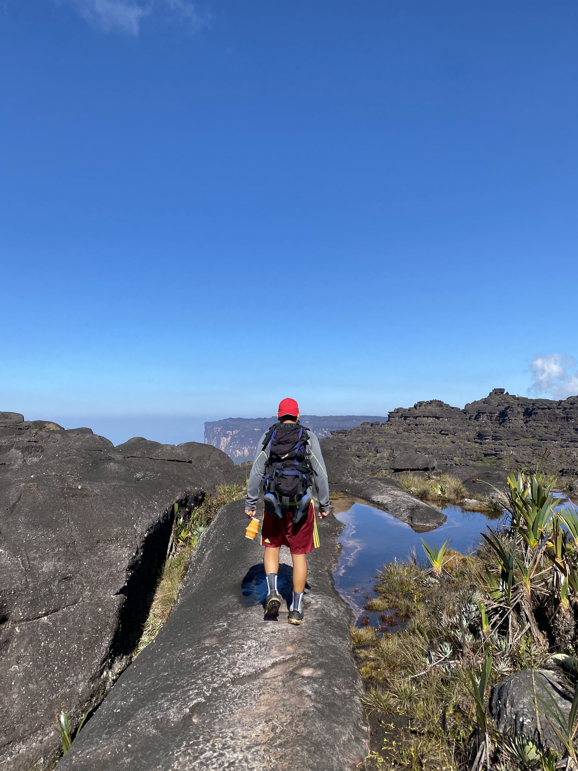 Hiker with a backpack walks on a rocky path towards a distant mountain, clear blue sky overhead.