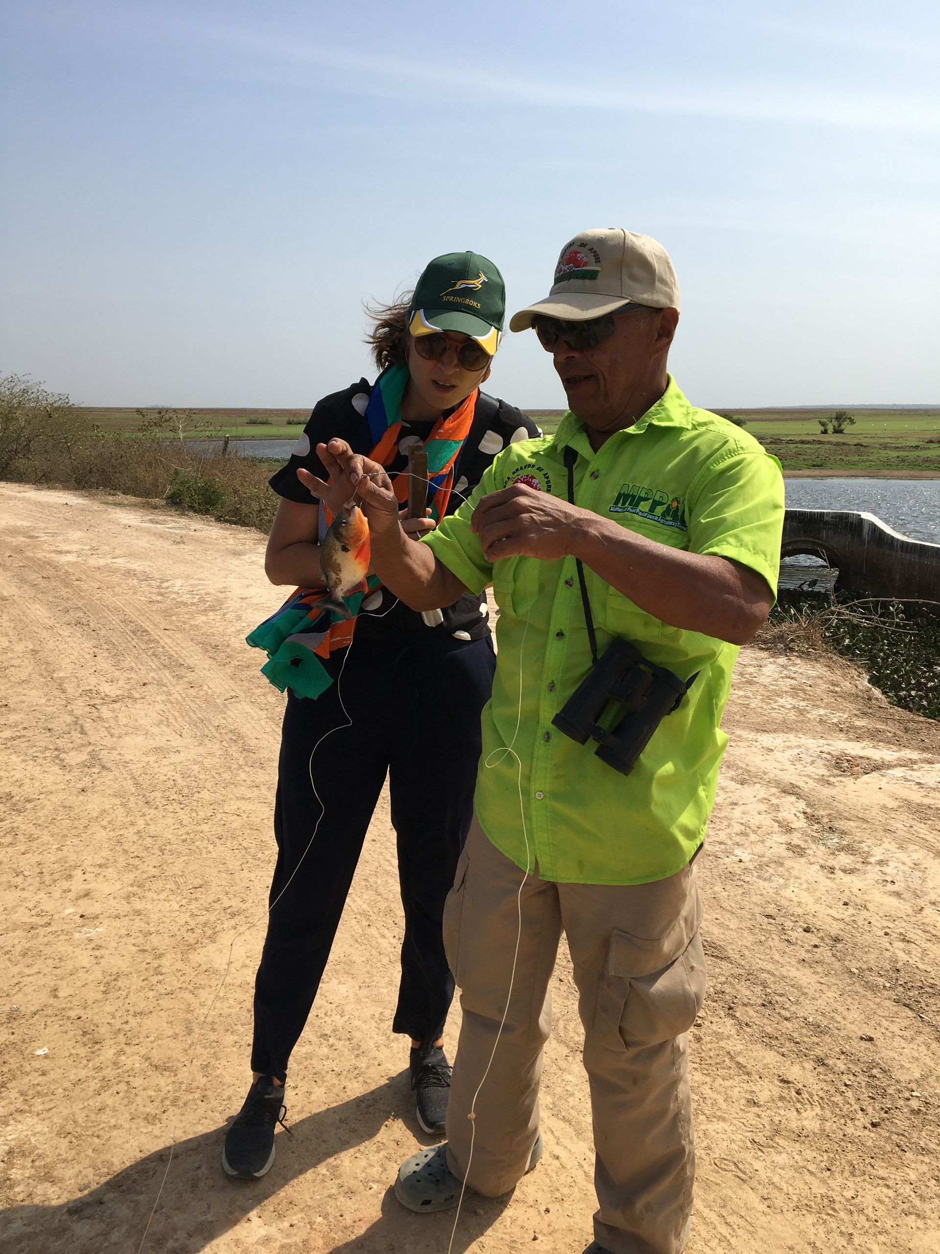 Two people outdoors, one in a lime shirt gestures towards the ground, explaining something to the woman in a baseball cap.