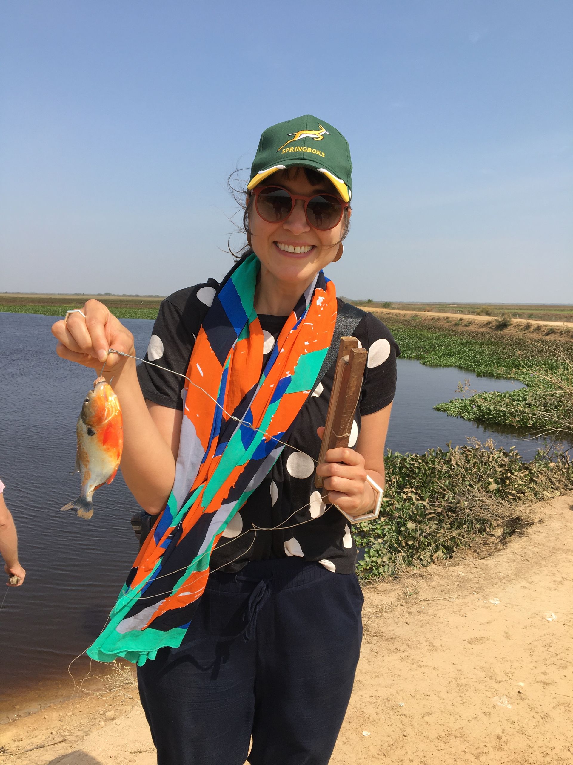Woman in hat and sunglasses holding a small orange fish and a wooden item, standing near a body of water.