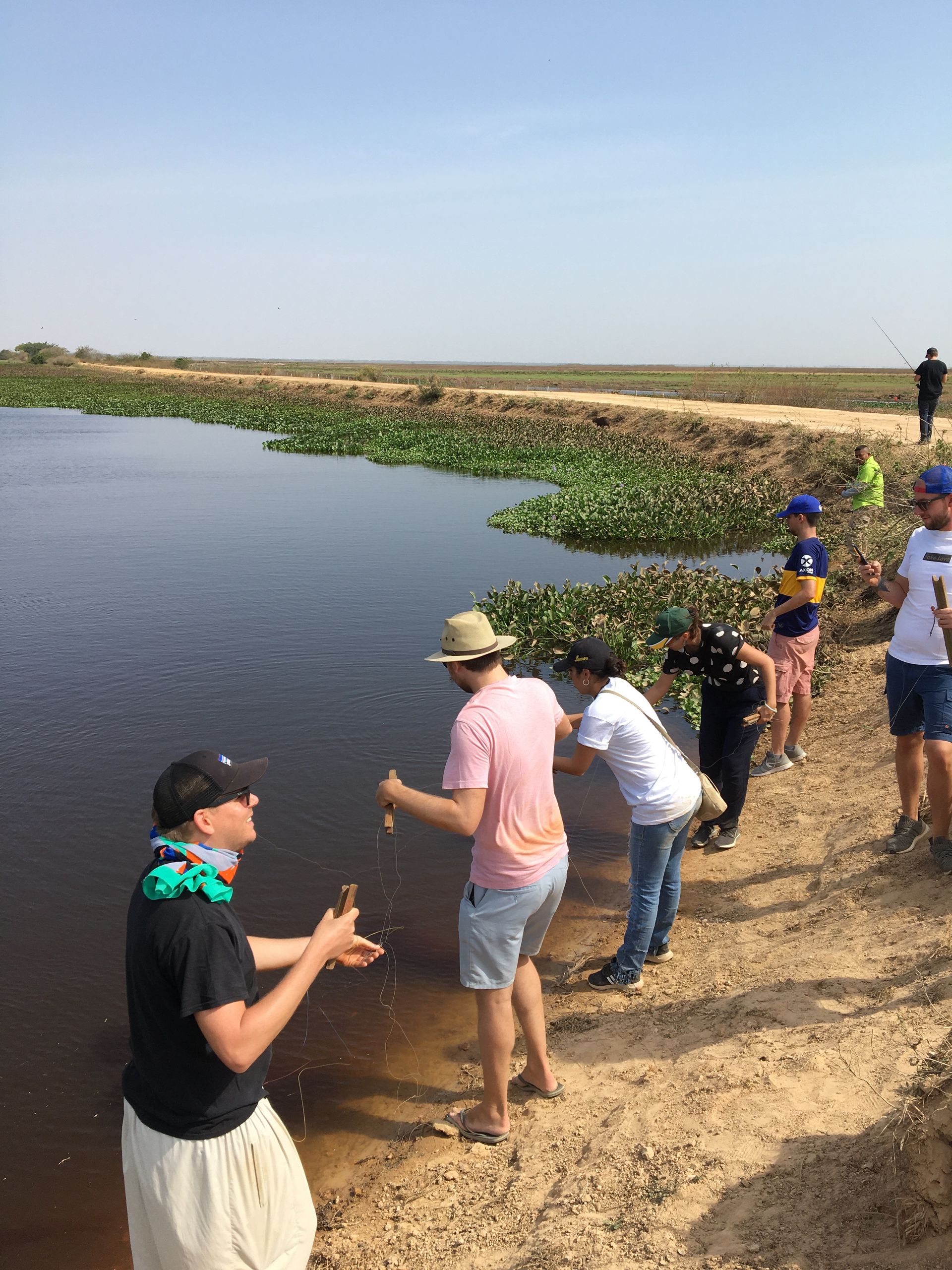 A group of people stands by a lake. One man pours something into the water, possibly a bottle. The setting is outdoors on a sunny day.