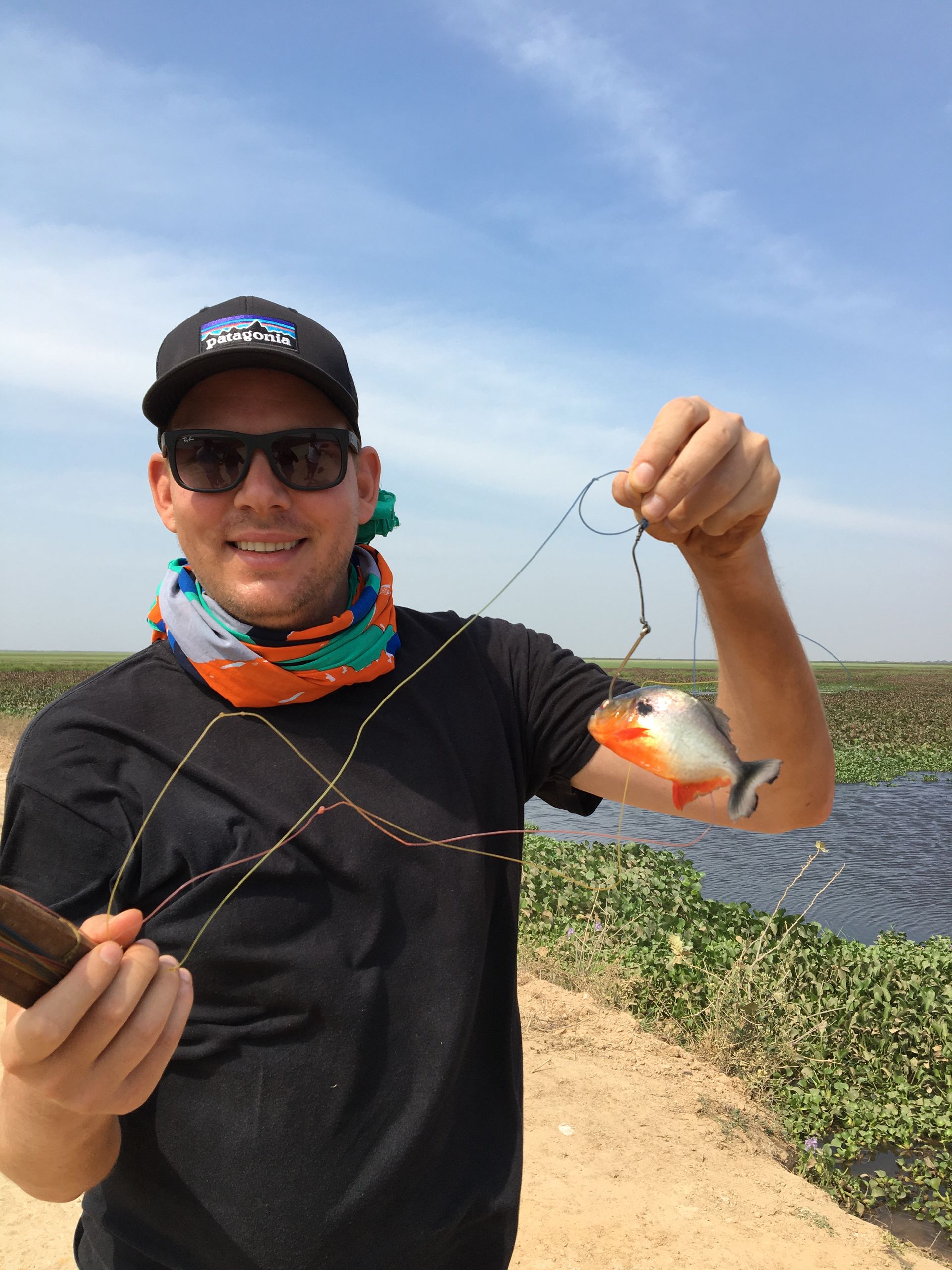 Man holding a small piranha fish by its mouth. He wears sunglasses, a black shirt, and a bandana. The setting is outdoors near a waterway.