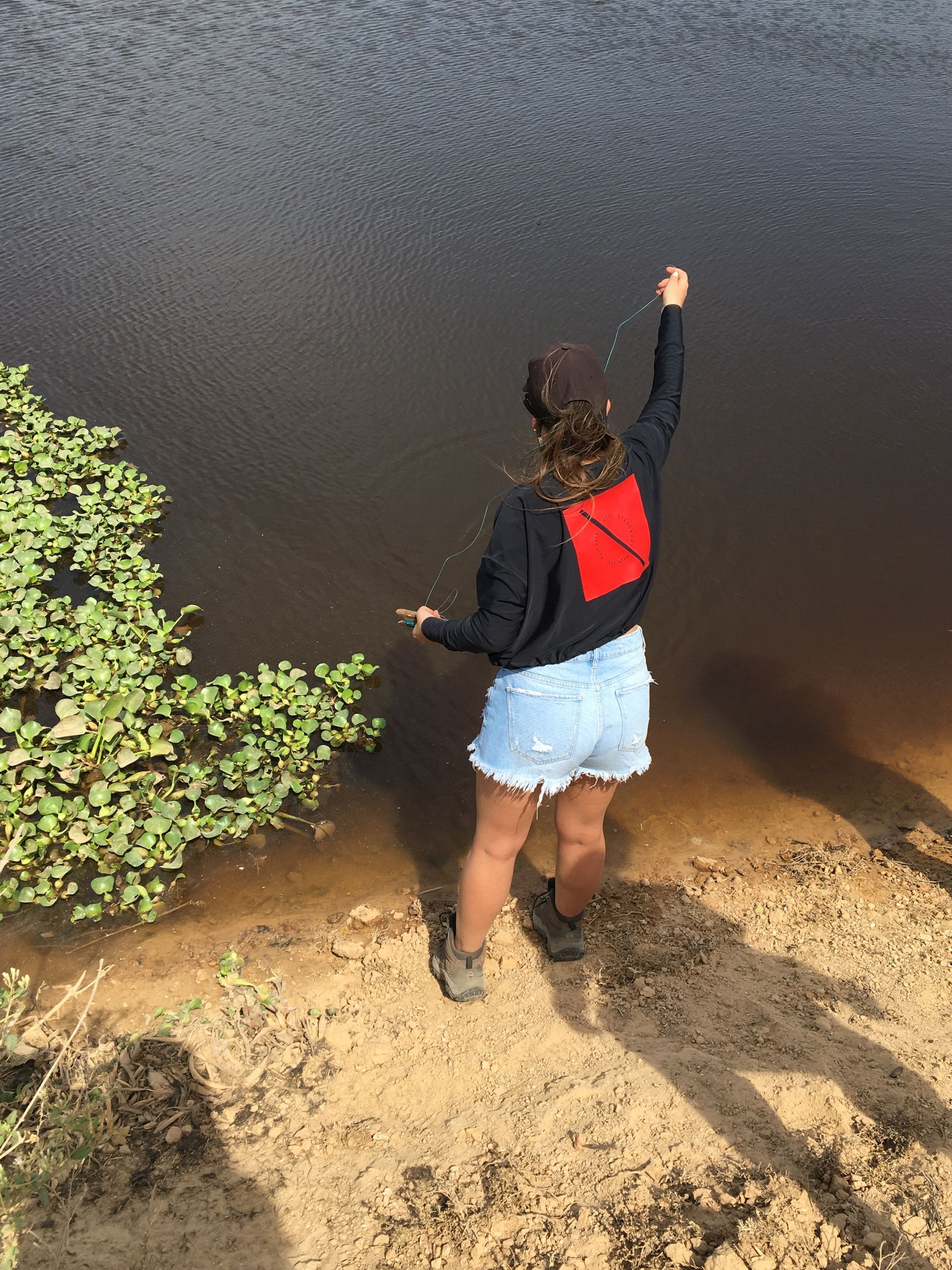 Woman fishing by a dark lake, wearing a black shirt with a red design, denim shorts, and boots.