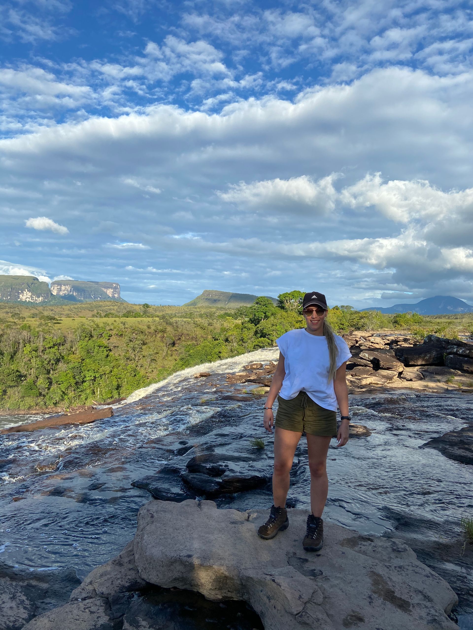 Woman in hat and shorts stands on a rocky ledge overlooking a lush green forest and mountains under a cloudy sky.