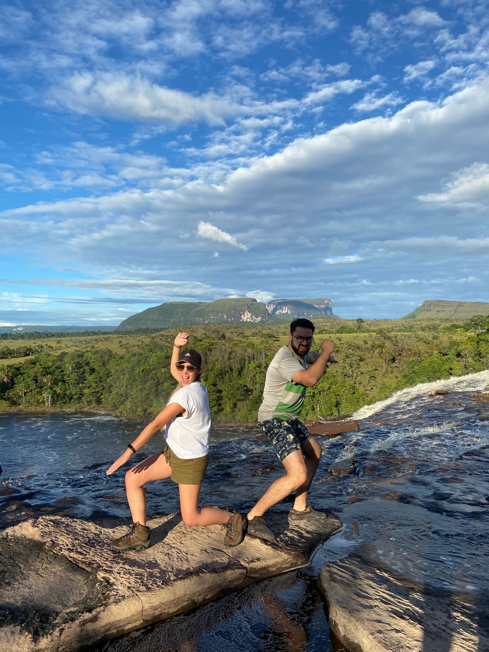 Two people posing playfully on a rocky ledge with a scenic mountain and sky backdrop. They appear to be celebrating.
