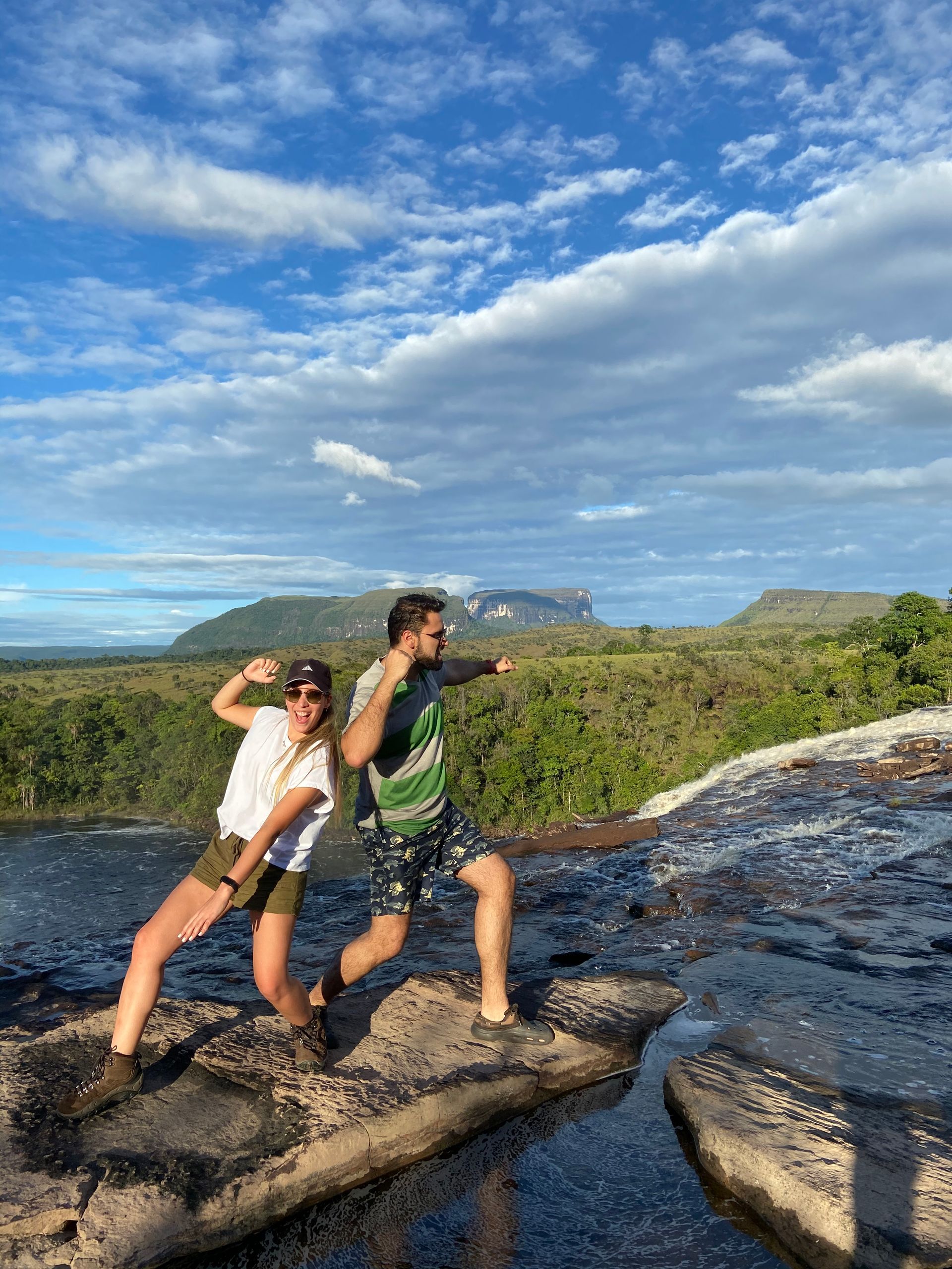Two people strike a pose on a rocky overlook with a scenic view of green hills and blue sky.