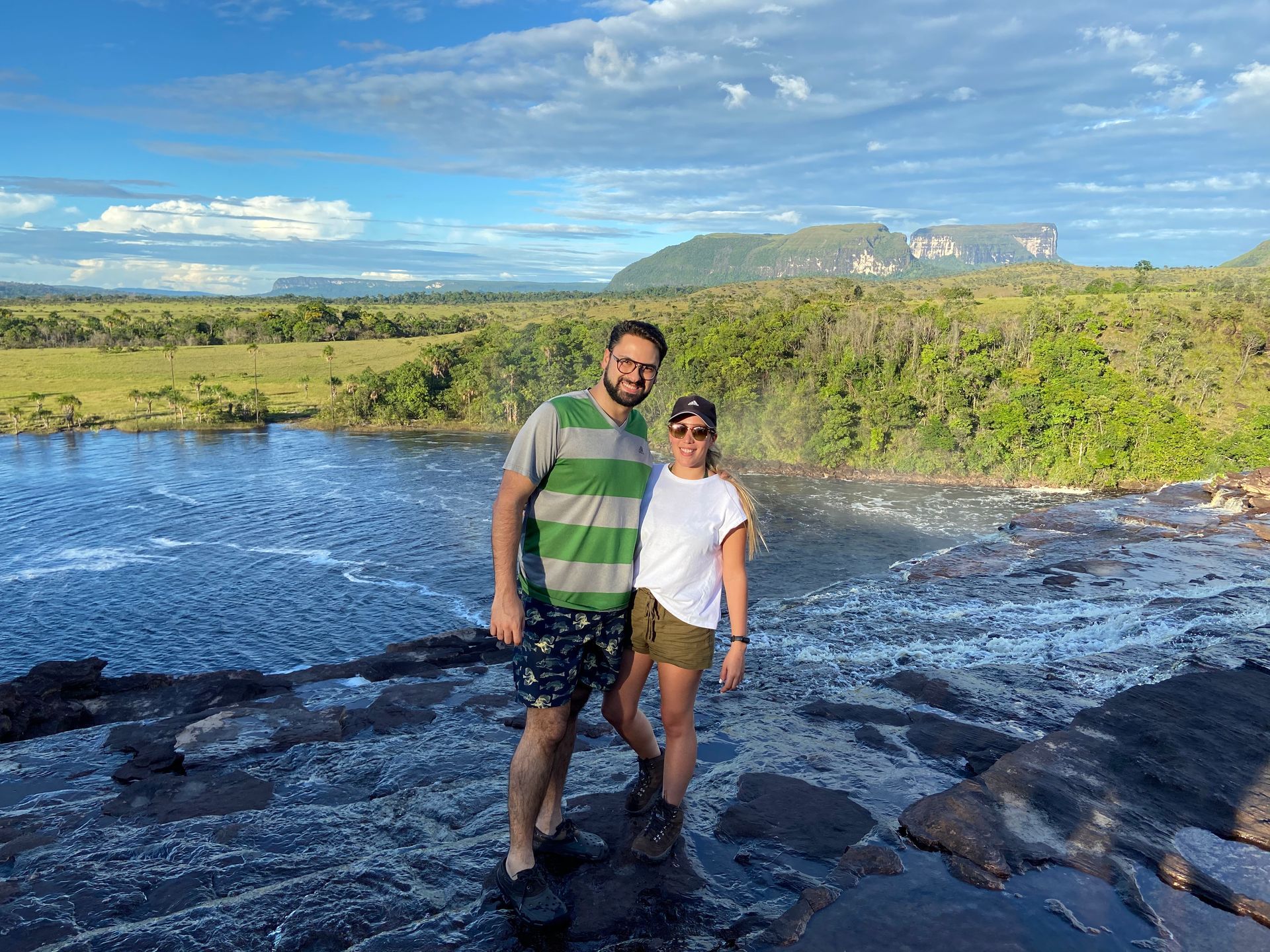 Couple stands on a rock ledge near a waterfall, with a grassy landscape and mountains in the background. They both are smiling.