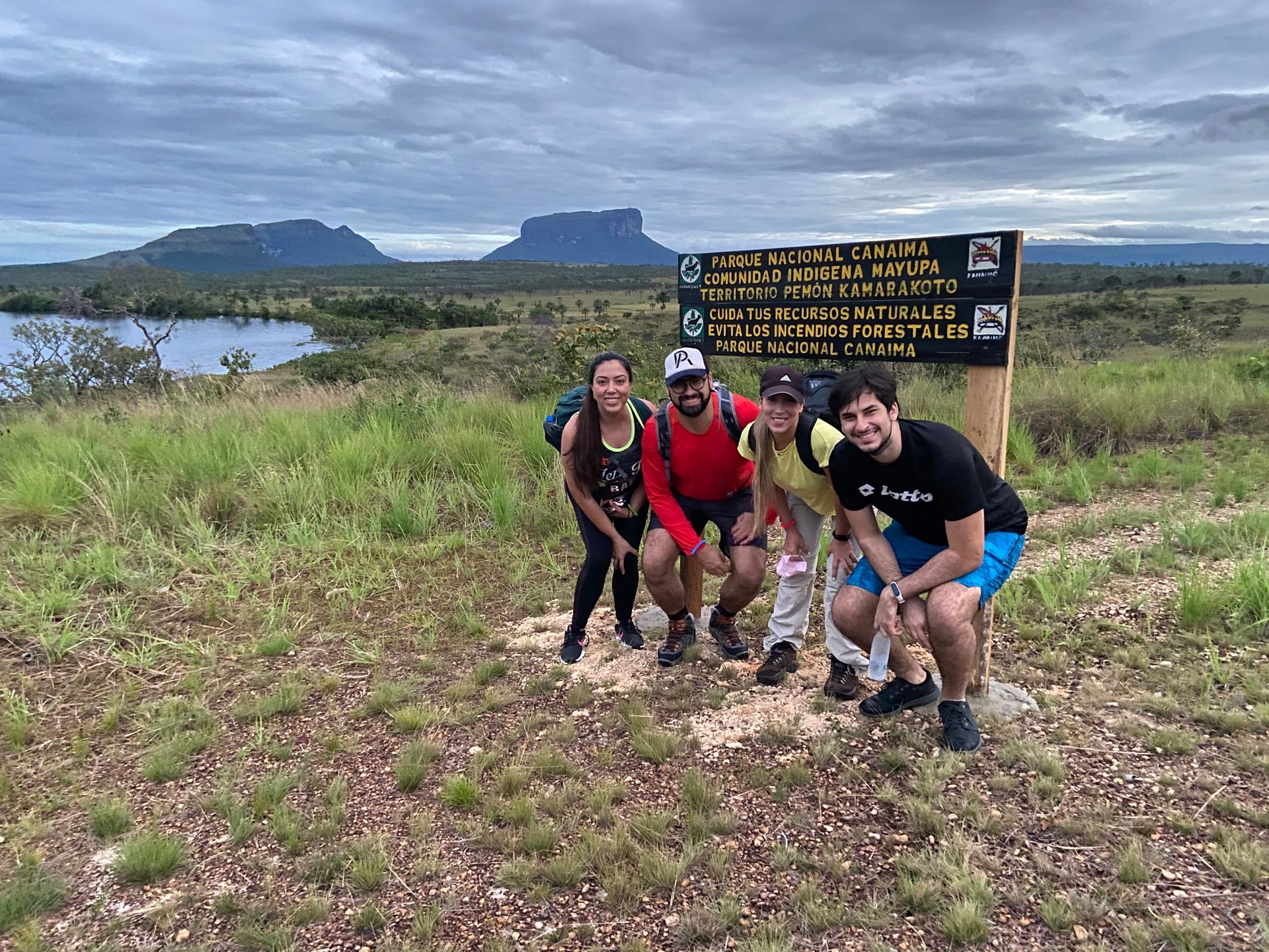 Group of five hikers pose by a sign in a grassy landscape with mountains and a lake under a cloudy sky.