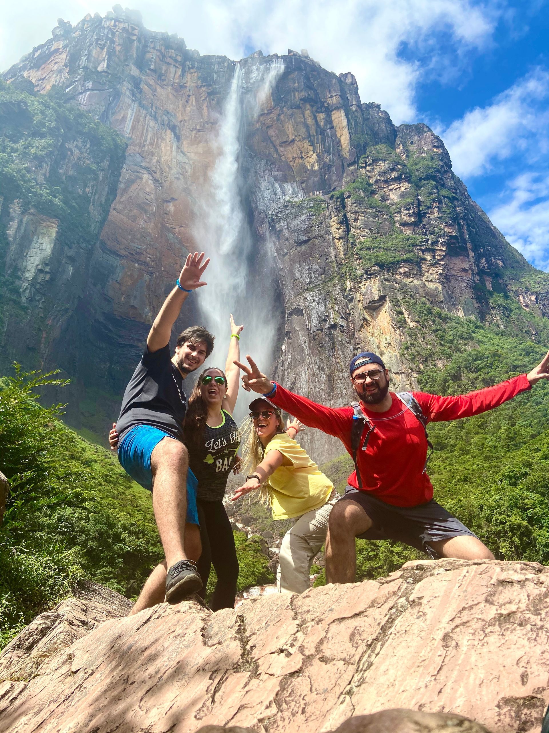 Group of four people pose with arms raised in front of a tall waterfall cascading down a rock face. Bright blue sky overhead.