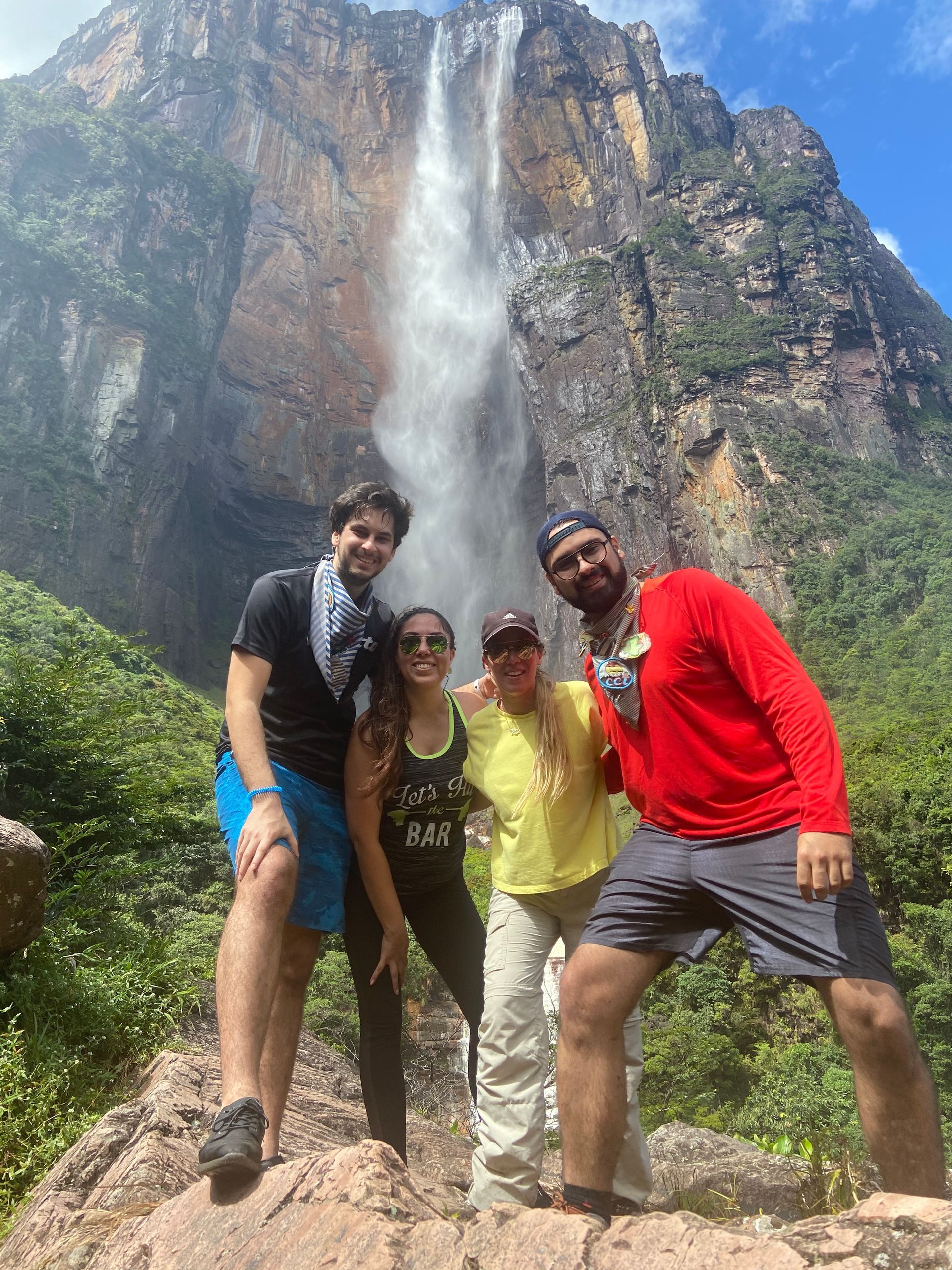 Four people pose in front of a tall waterfall. Green foliage surrounds them; the sky is blue.
