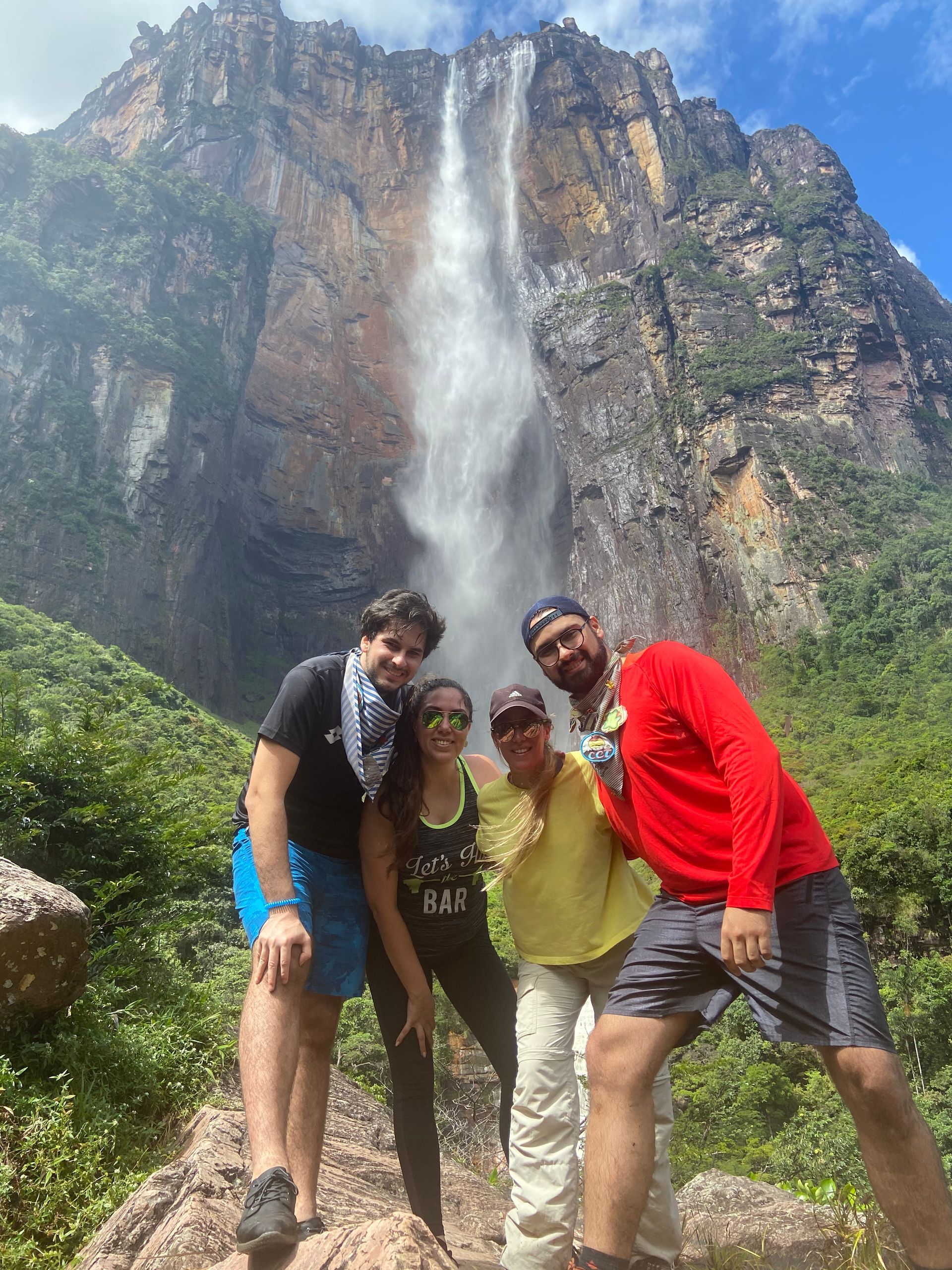 Four people pose in front of Angel Falls, Venezuela. They stand on a path, smiling at the camera against a backdrop of the massive waterfall.