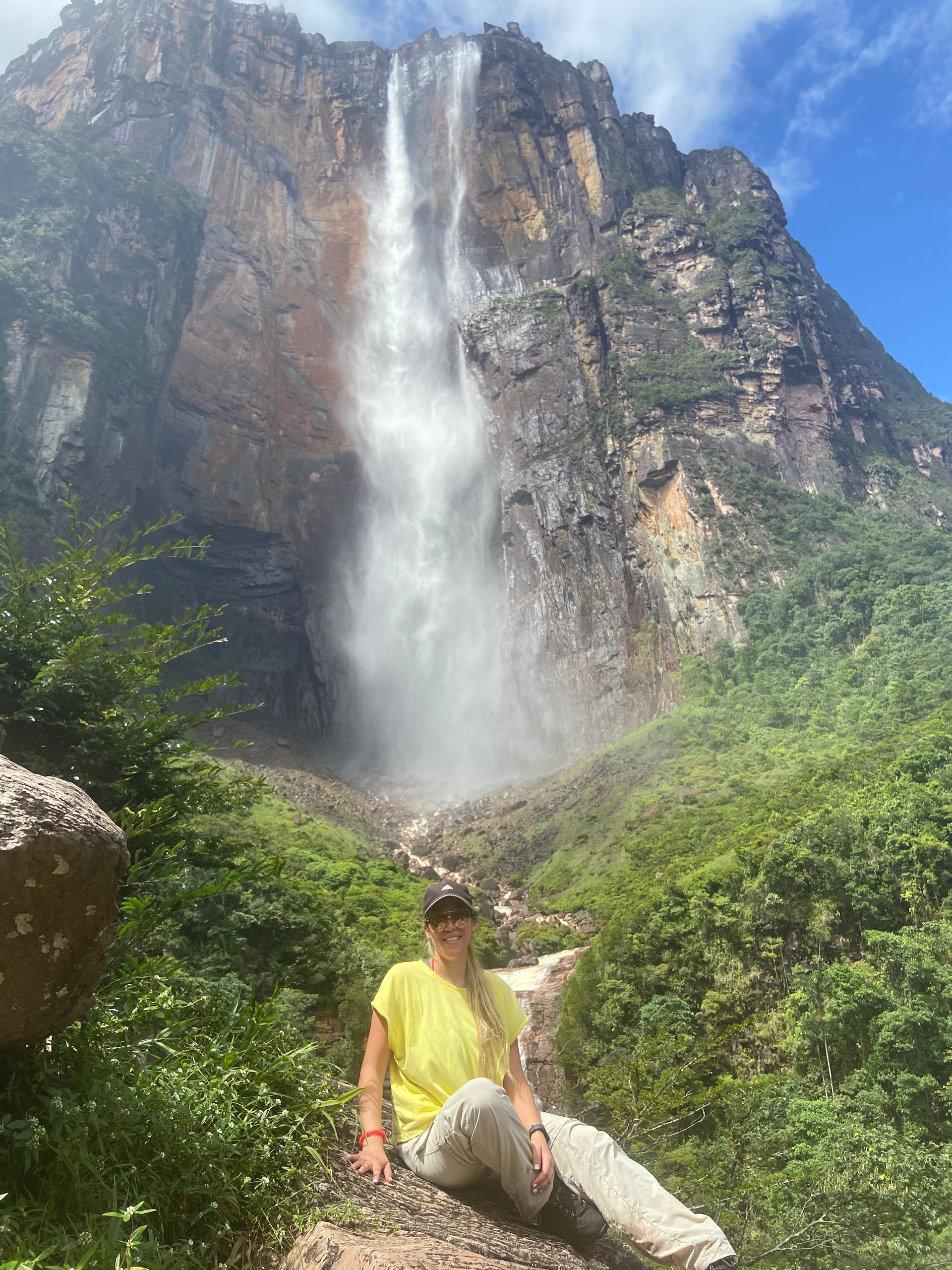 Woman in yellow shirt and beige pants sits before Angel Falls in Venezuela. Lush green vegetation surrounds her.