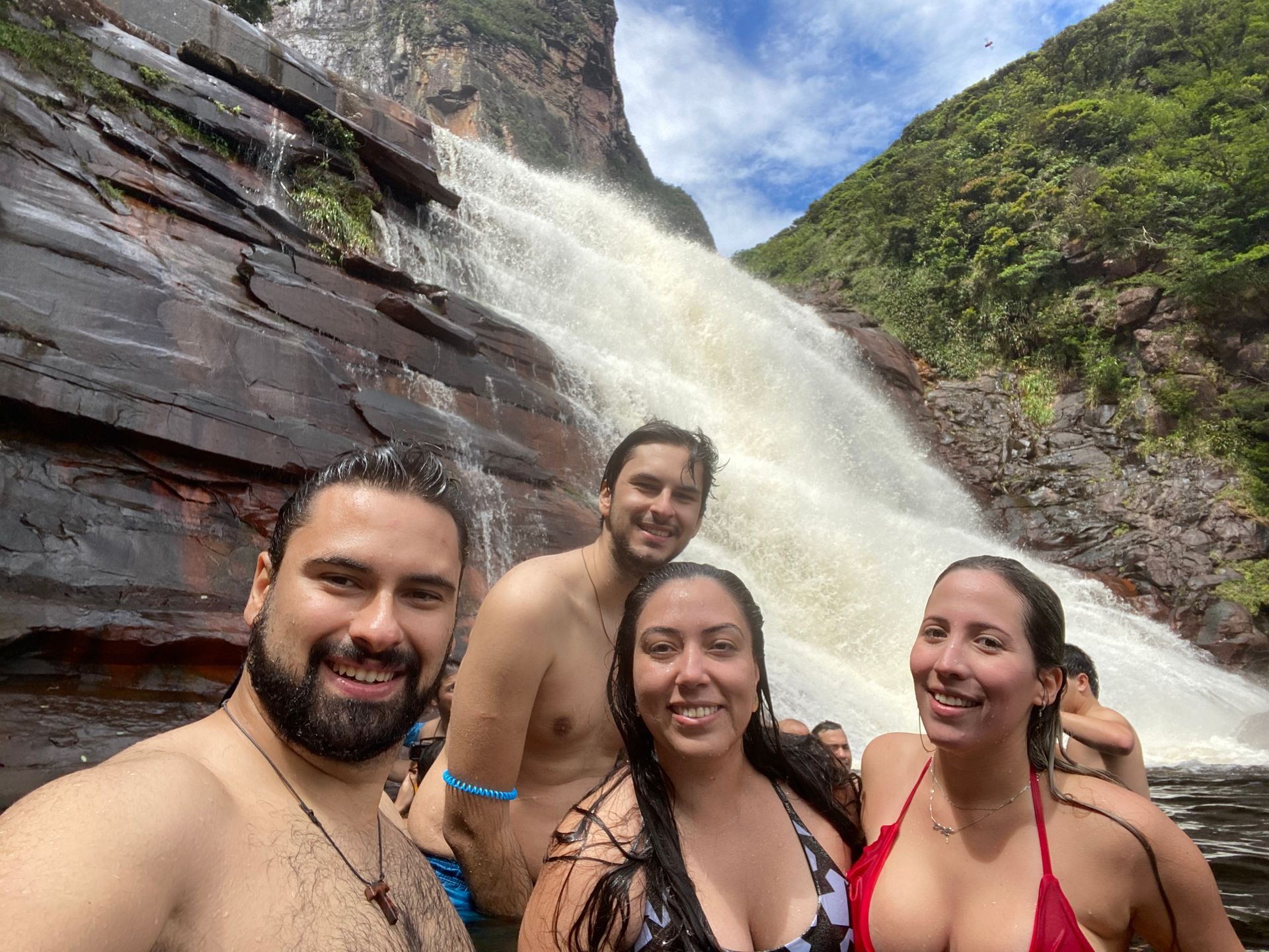 Four people smiling in front of a waterfall, enjoying the water. Dark rocks surround them, with lush green vegetation to the side.