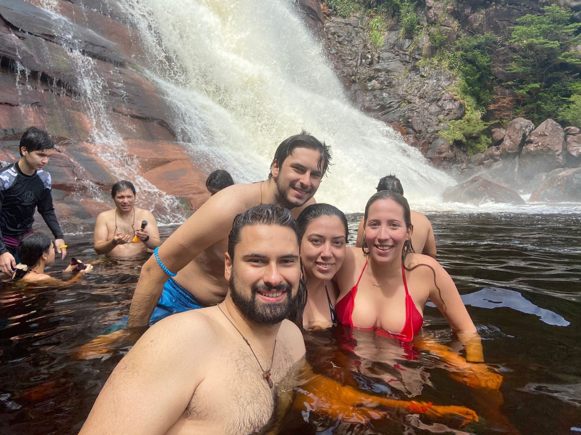 Group of people smiling in a dark-colored pool at the base of a waterfall, with a red-bikini-clad woman prominent.