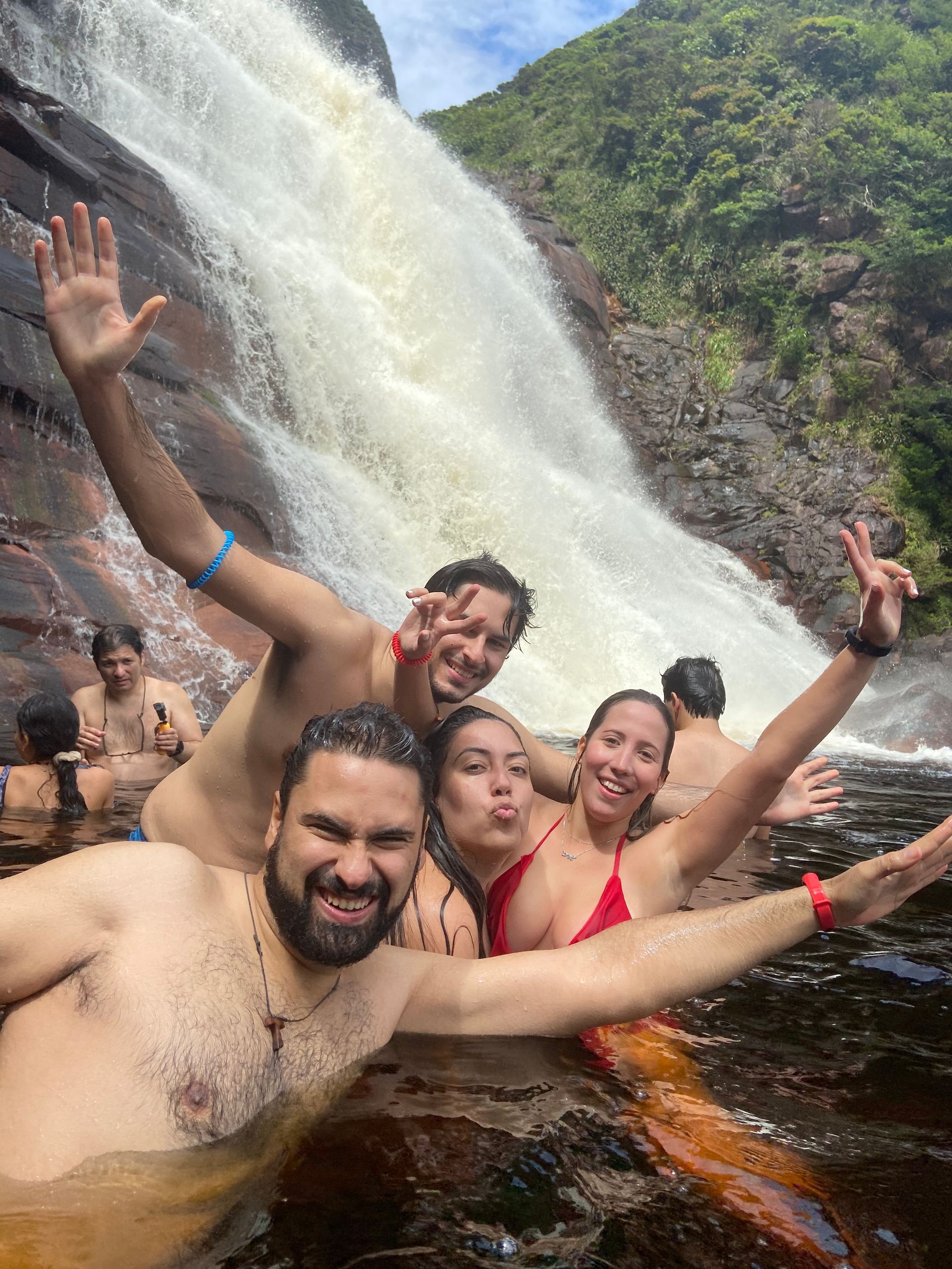 Group of people enjoying a waterfall. Some are swimming, smiling, and waving in the water. Lush green foliage surrounds.