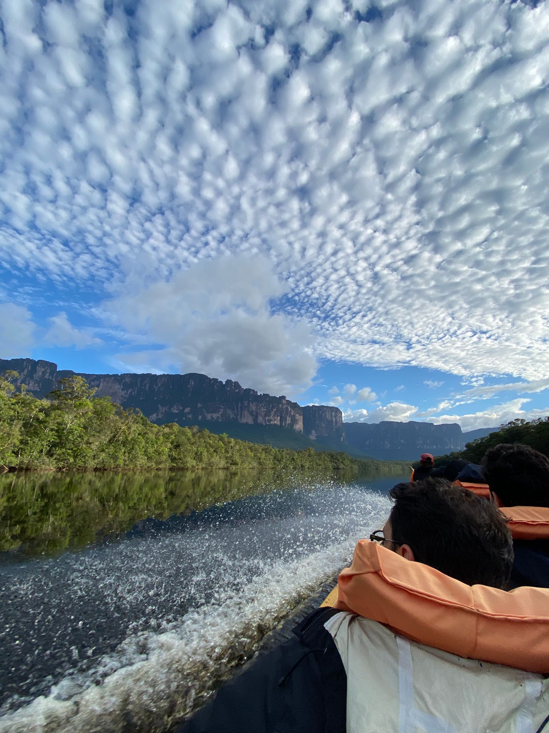 Boat on a calm lake with distant mountains under a sky filled with fluffy white clouds. Passengers wear orange life vests.