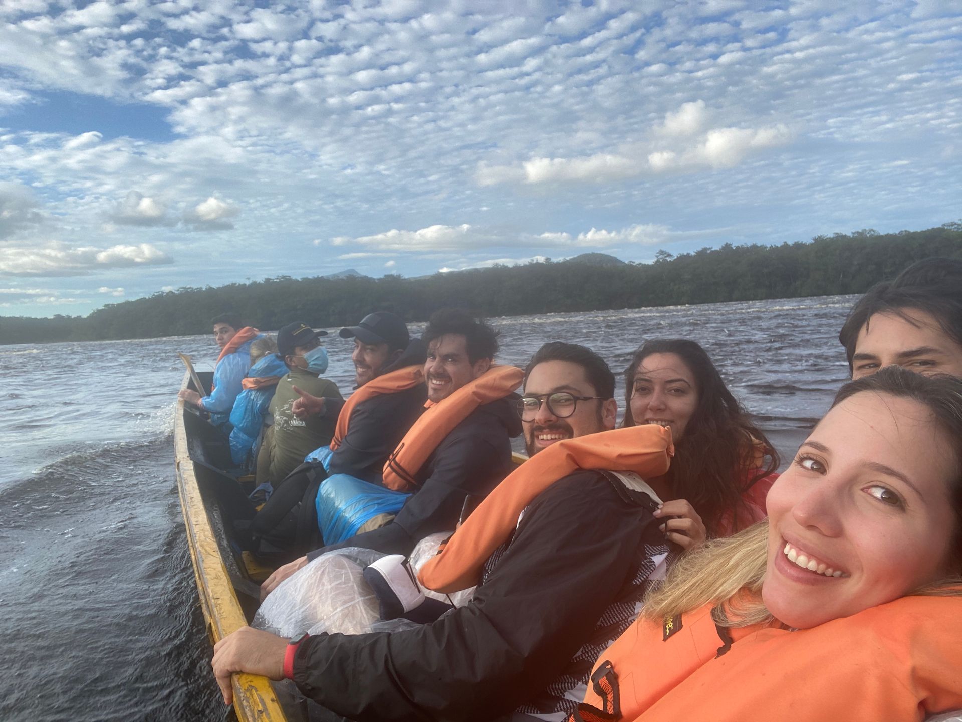 A group of people wearing orange life vests smile while riding in a long wooden boat on a lake, under a cloudy sky.