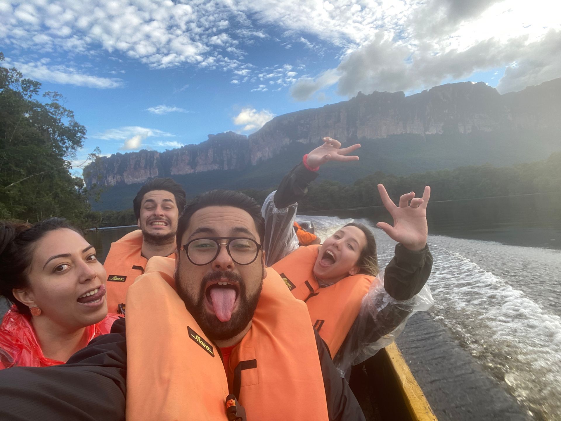 Four people in orange life vests on a boat, smiling and making playful gestures. A large mountain is visible in the background.