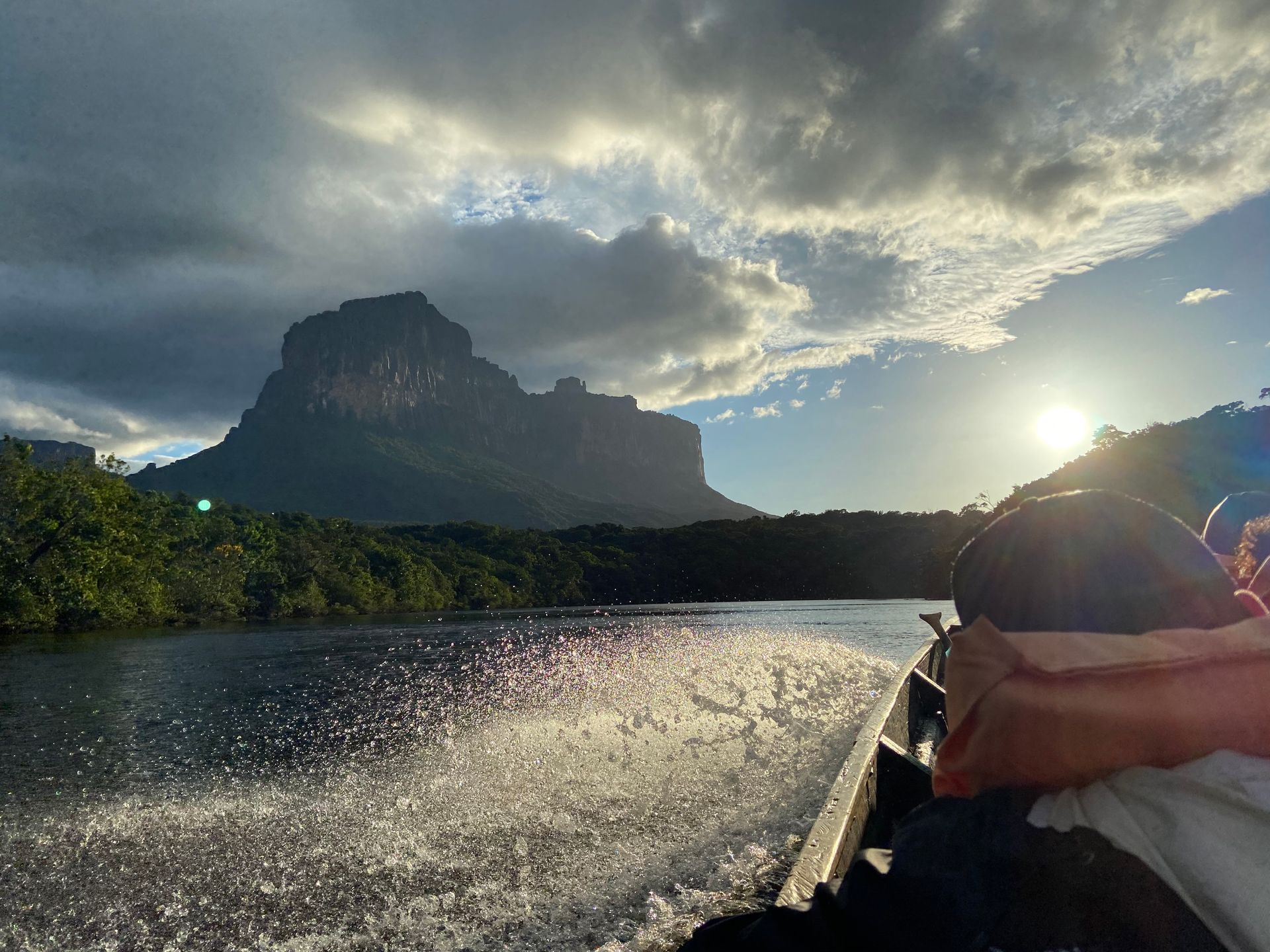 Boat on a dark river with spray, approaching a large mountain silhouetted by the sun setting behind cloudy skies.