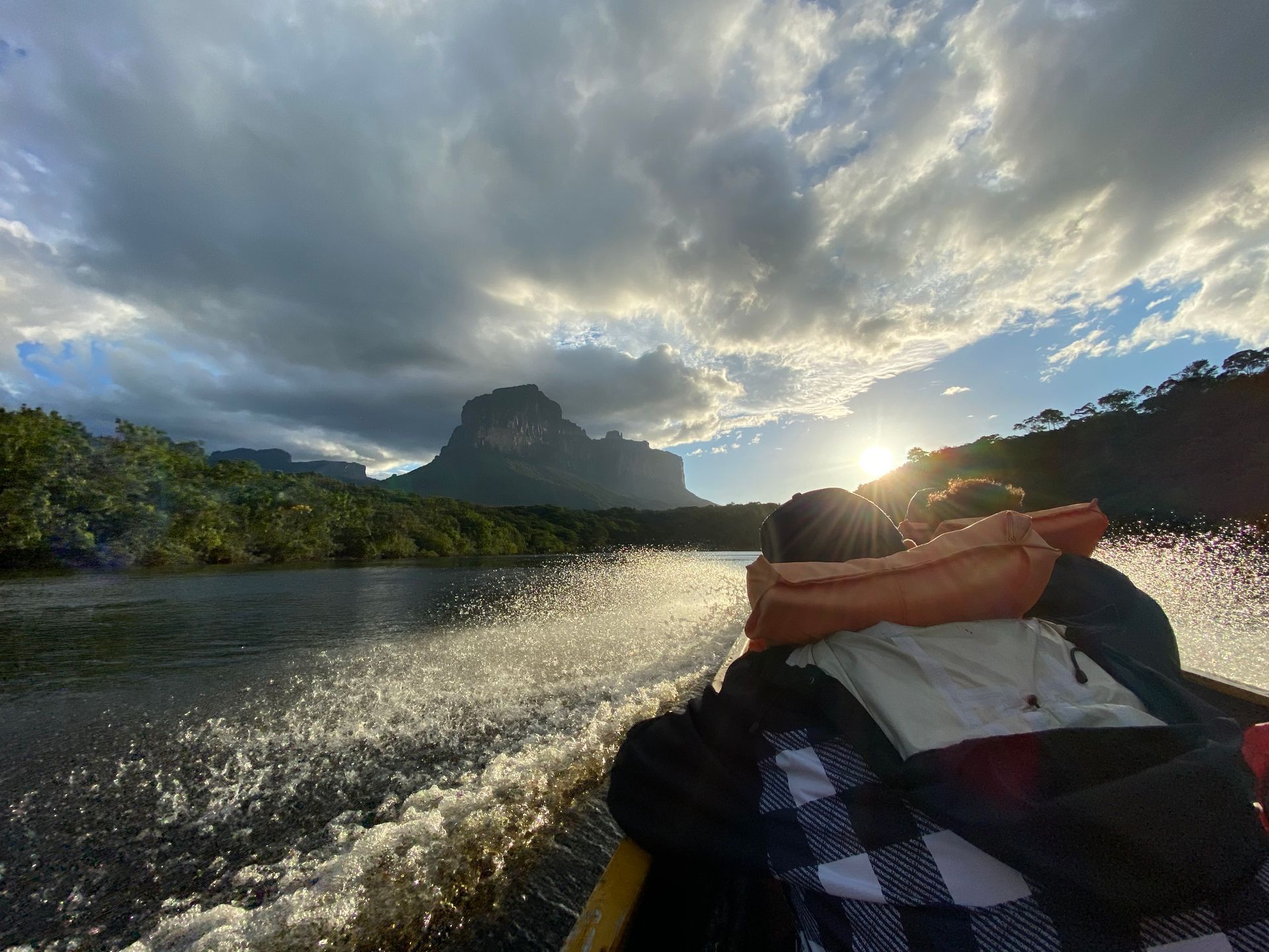 Person on boat looking at mountain and sunset over river under cloudy sky.