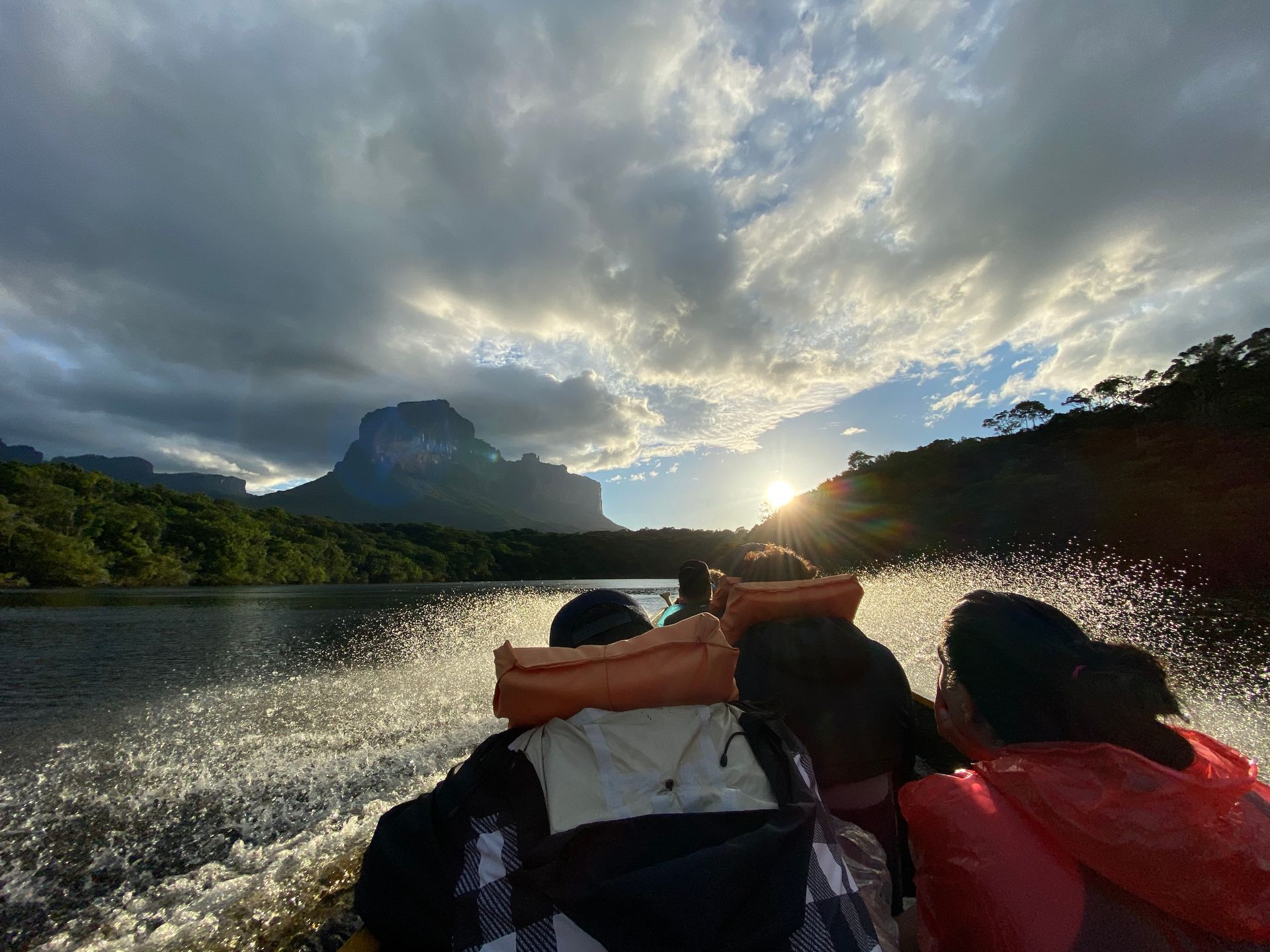 People in a boat on a lake, heading toward a sunset and a mountain in the distance. Water splashes around the boat.
