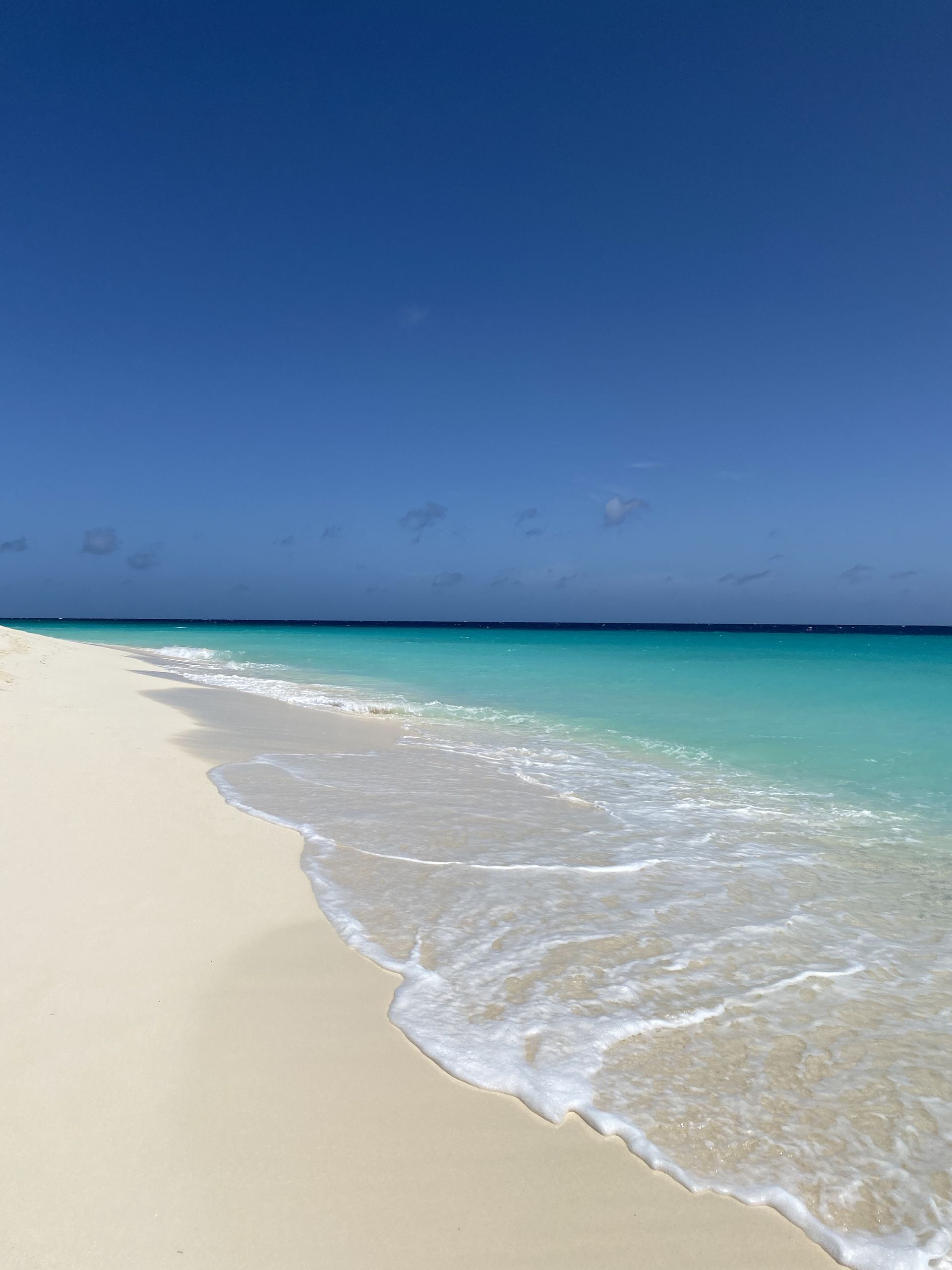 White sandy beach with crystal-clear turquoise water under a bright blue sky. Gentle waves lap the shore.