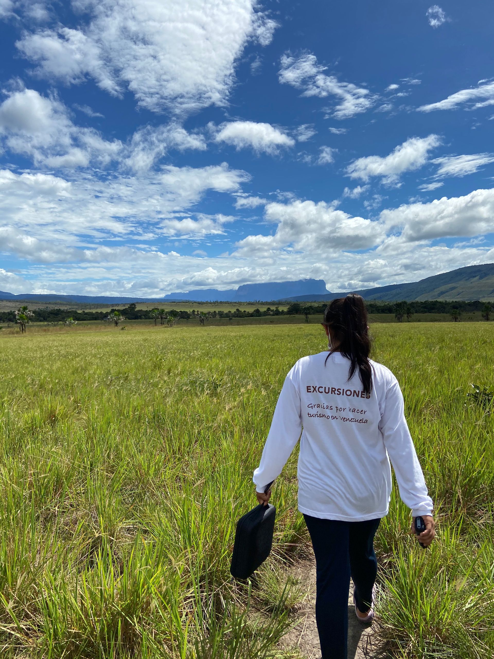 Woman walking through a grassy field towards mountains under a blue sky with fluffy clouds. She is holding a black bag and wearing a white shirt.