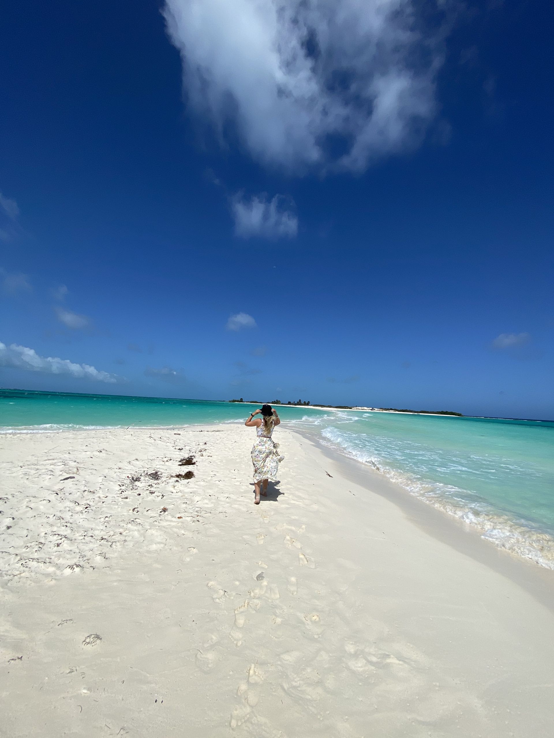 Woman walks on a white sandbar between turquoise water under a bright blue sky with fluffy clouds.