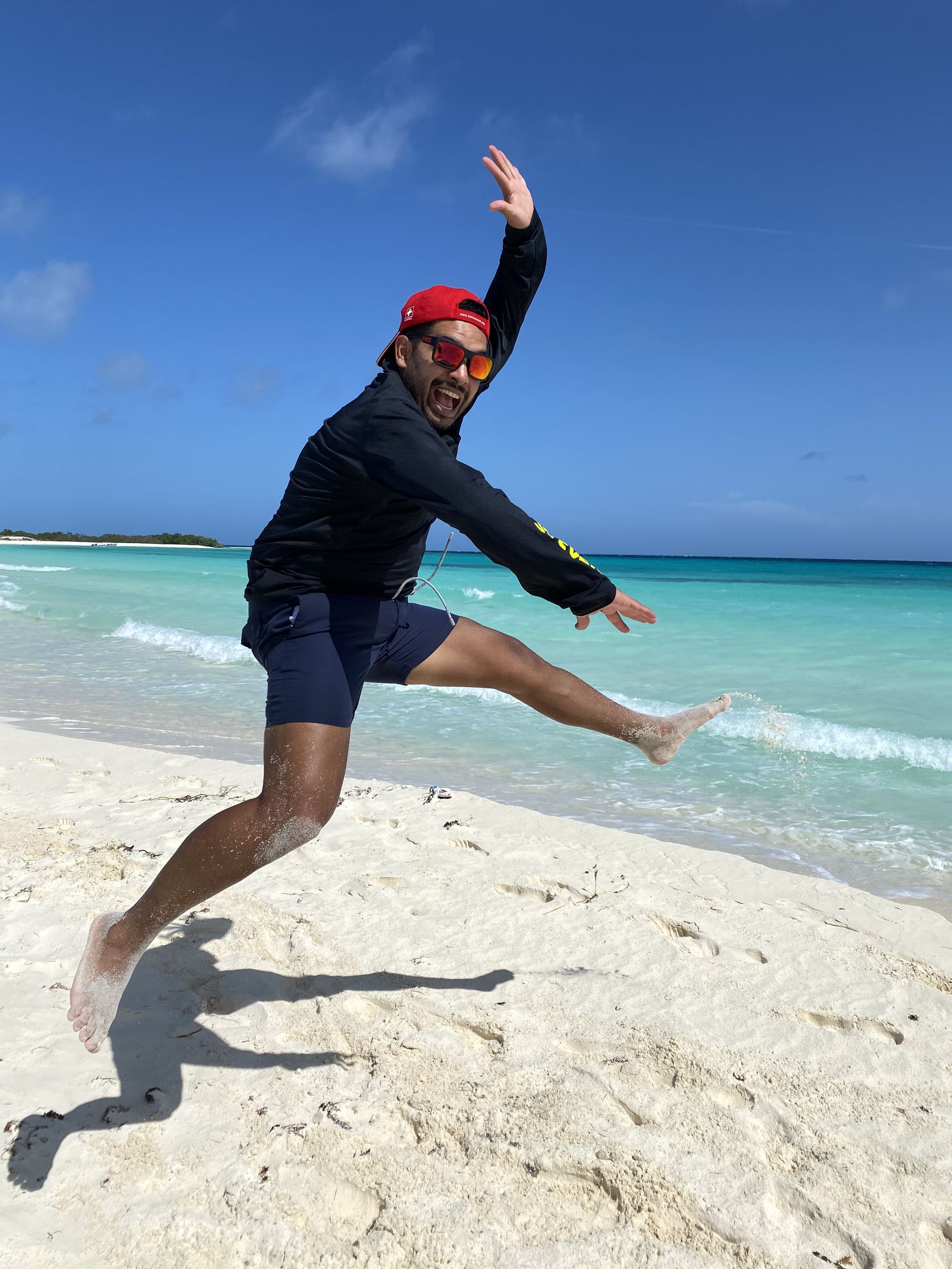 Man jumping on a beach with clear turquoise water and blue sky. He wears a red cap, sunglasses, and a black jacket.