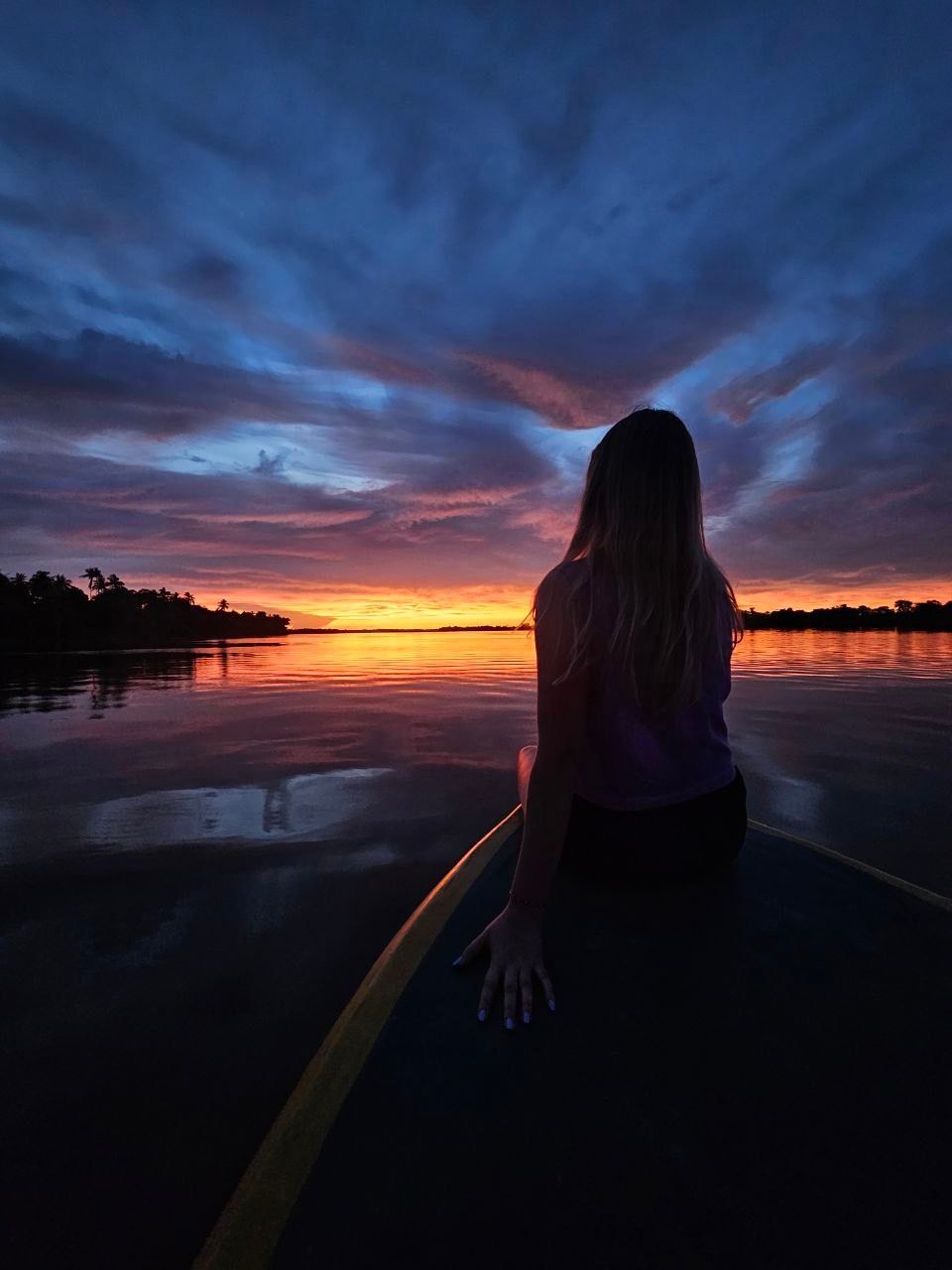 Woman sits on a board, facing a vibrant sunset over a calm body of water. Orange, pink, and blue hues fill the sky.