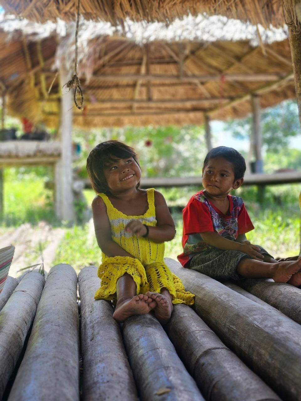 Two children sit on a wooden platform under a thatched roof. The child on the left smiles in a yellow dress; the other in red.