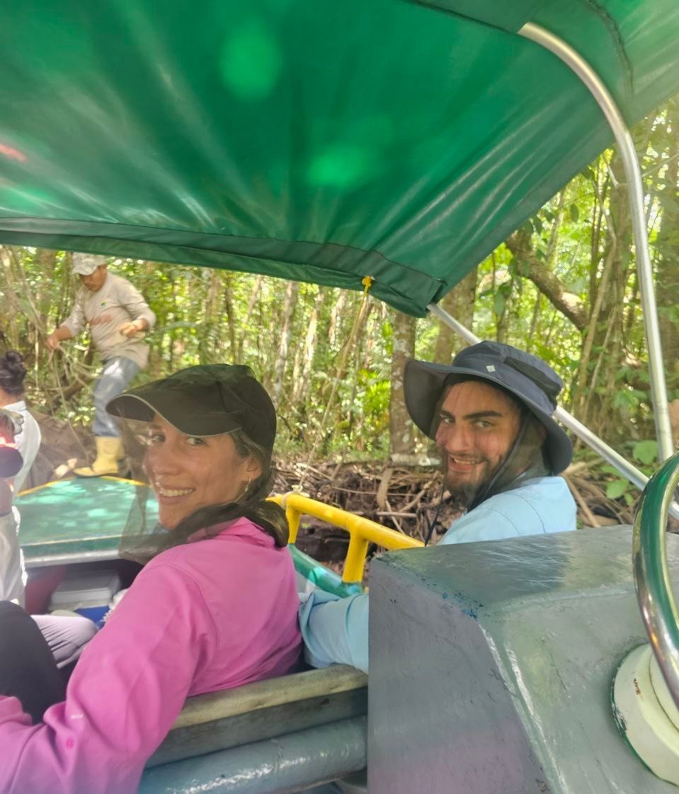 Two people ride in a small boat under a green canopy, smiling at the camera. They are in a lush, green forest setting.