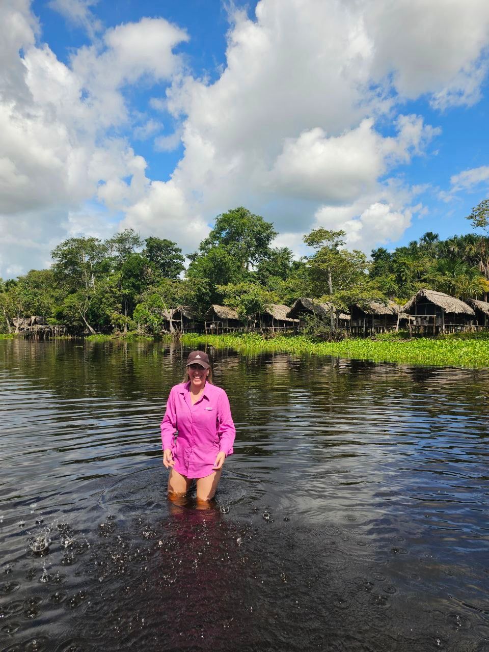 Woman in pink shirt wading in dark water; thatched-roof huts and lush green trees line the shore under a partly cloudy sky.