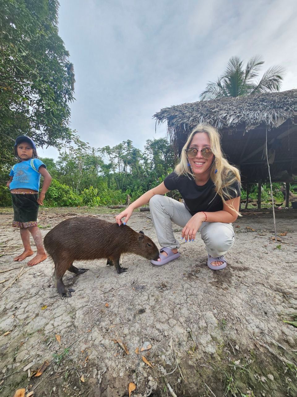 Woman petting a capybara, child watching. Outdoors by a thatched roof shelter; neutral tones.