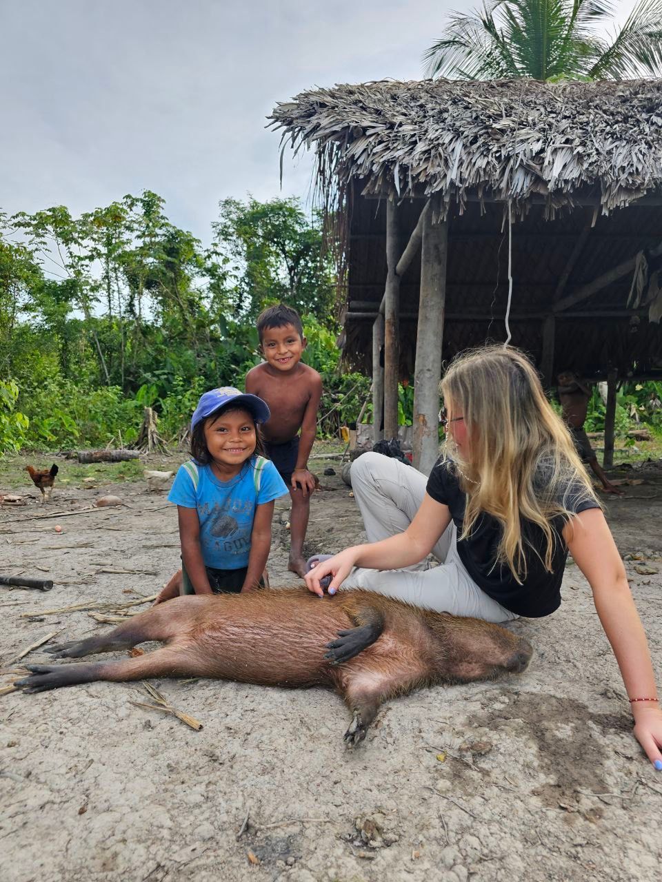 A fair-skinned person pets a large, dead animal on the ground as two young children watch in front of a thatched-roof structure in a rural setting.