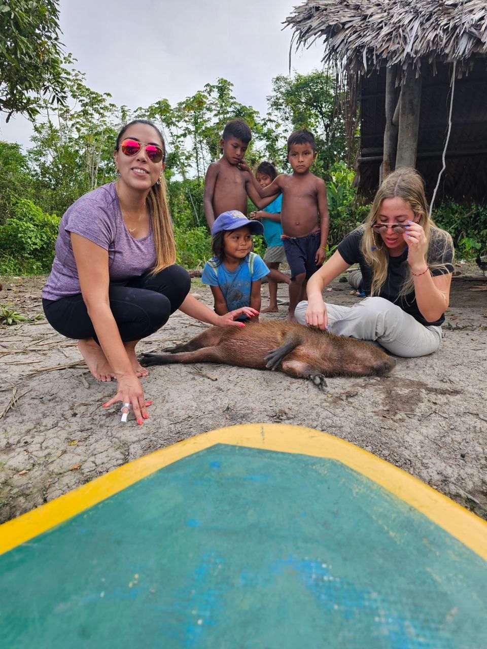 Two women touch a capybara on the ground as children watch near a boat and a thatched hut. The scene takes place outdoors.