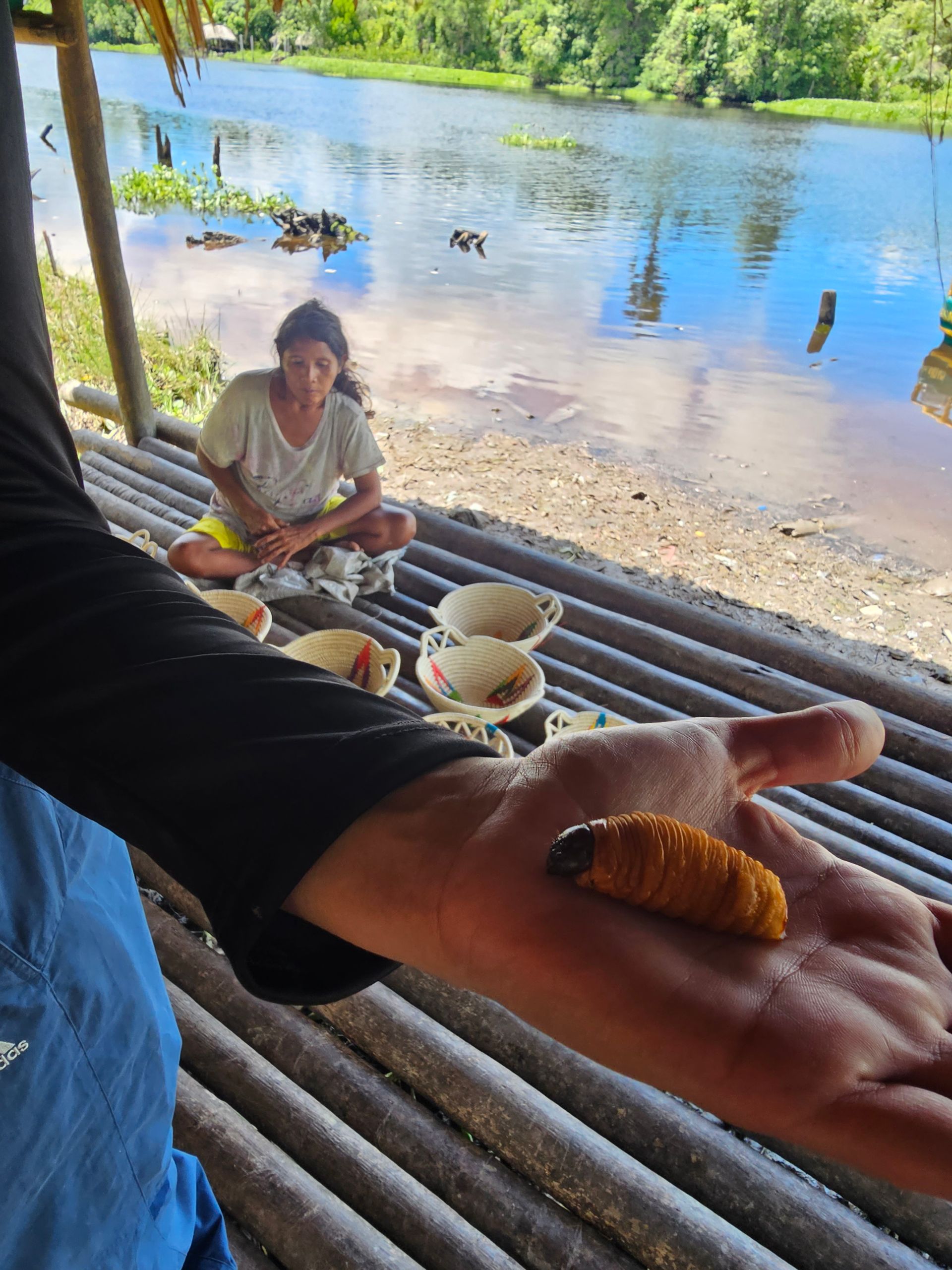A person holds a large, grub-like insect in their palm. Another person sits nearby, with bowls, by a river.