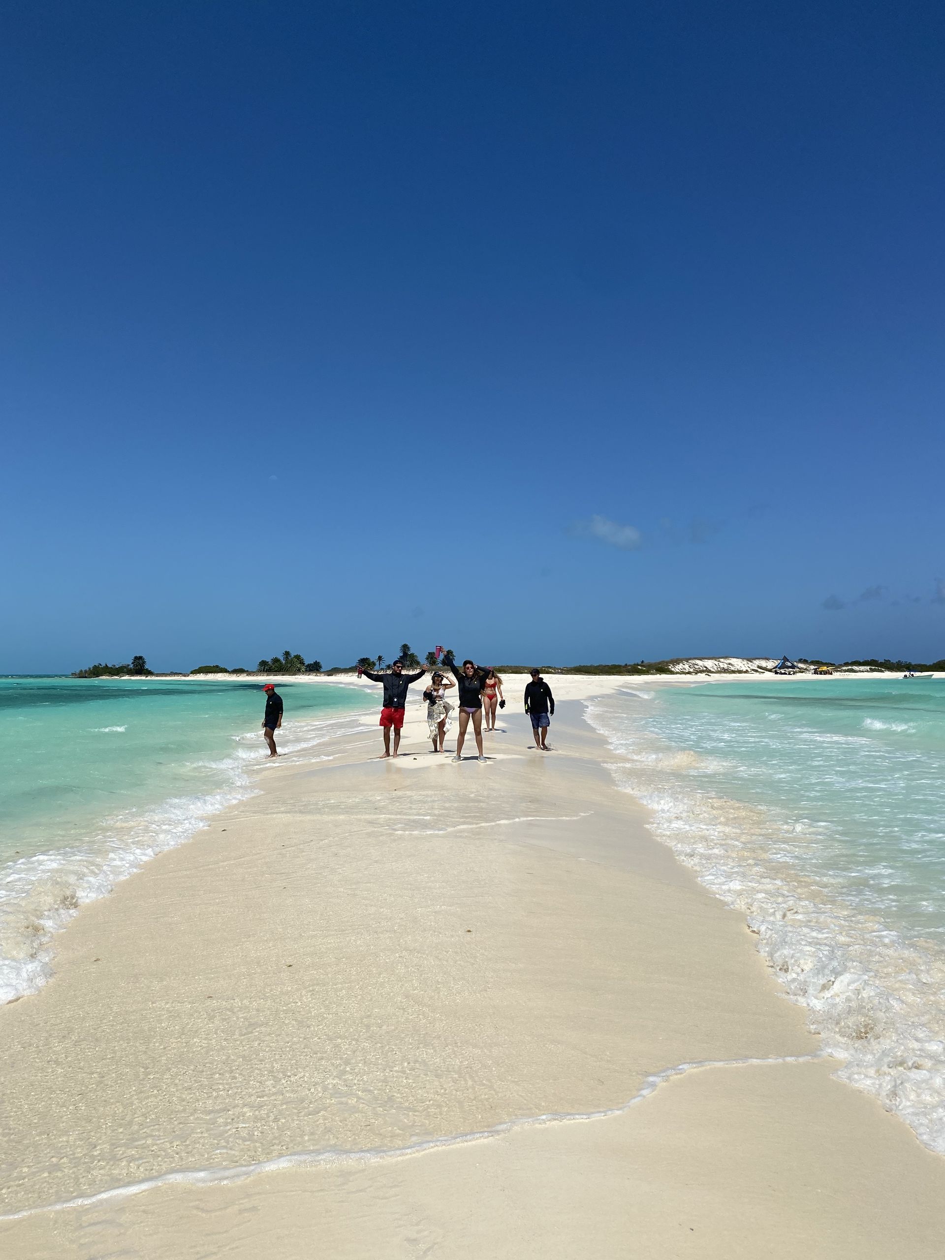 People walking on a sandbar in shallow turquoise water under a bright blue sky.