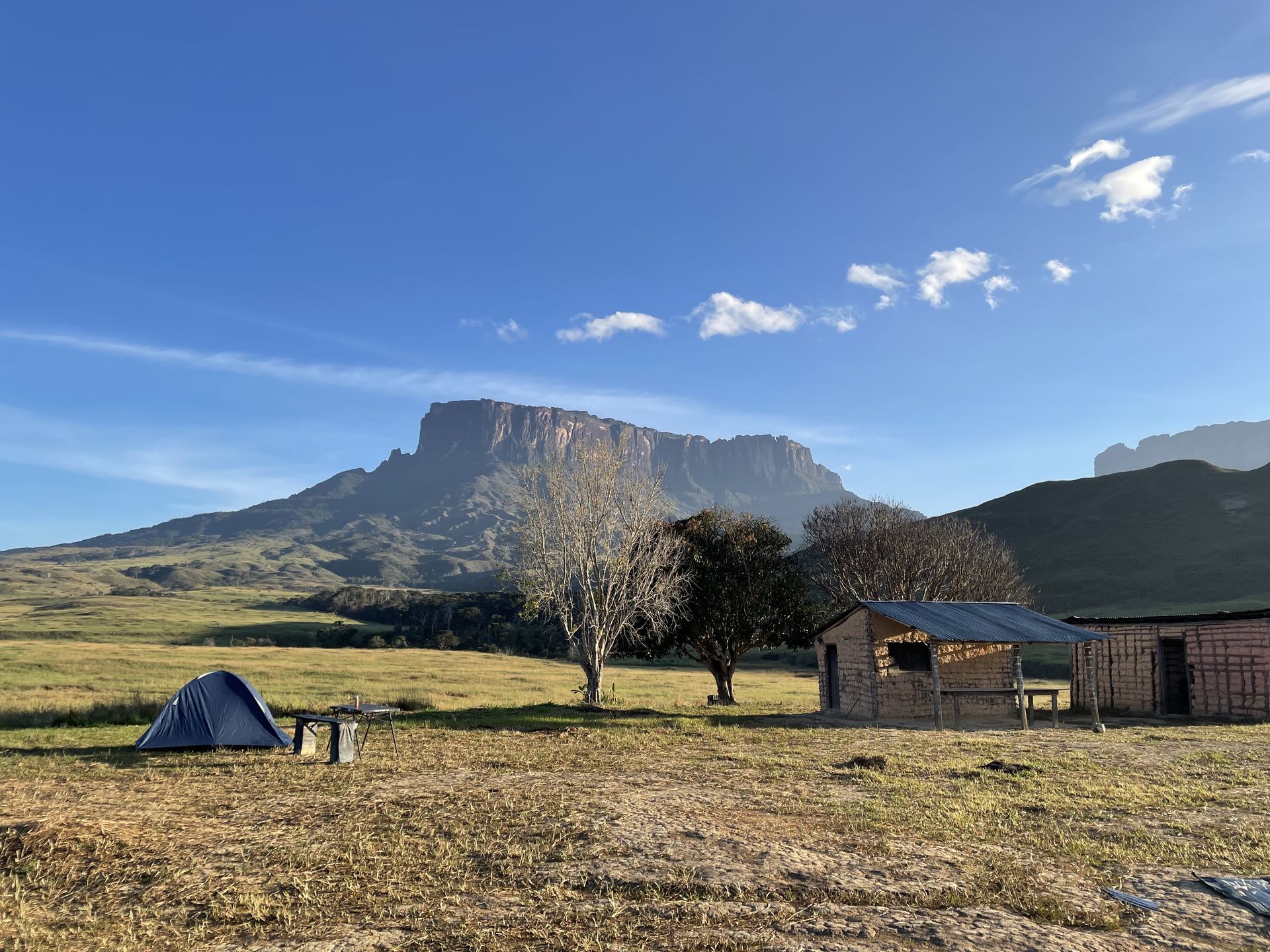 A campsite in a grassy field with a tent, rustic cabins, and a large flat-topped mountain in the background under a blue sky.