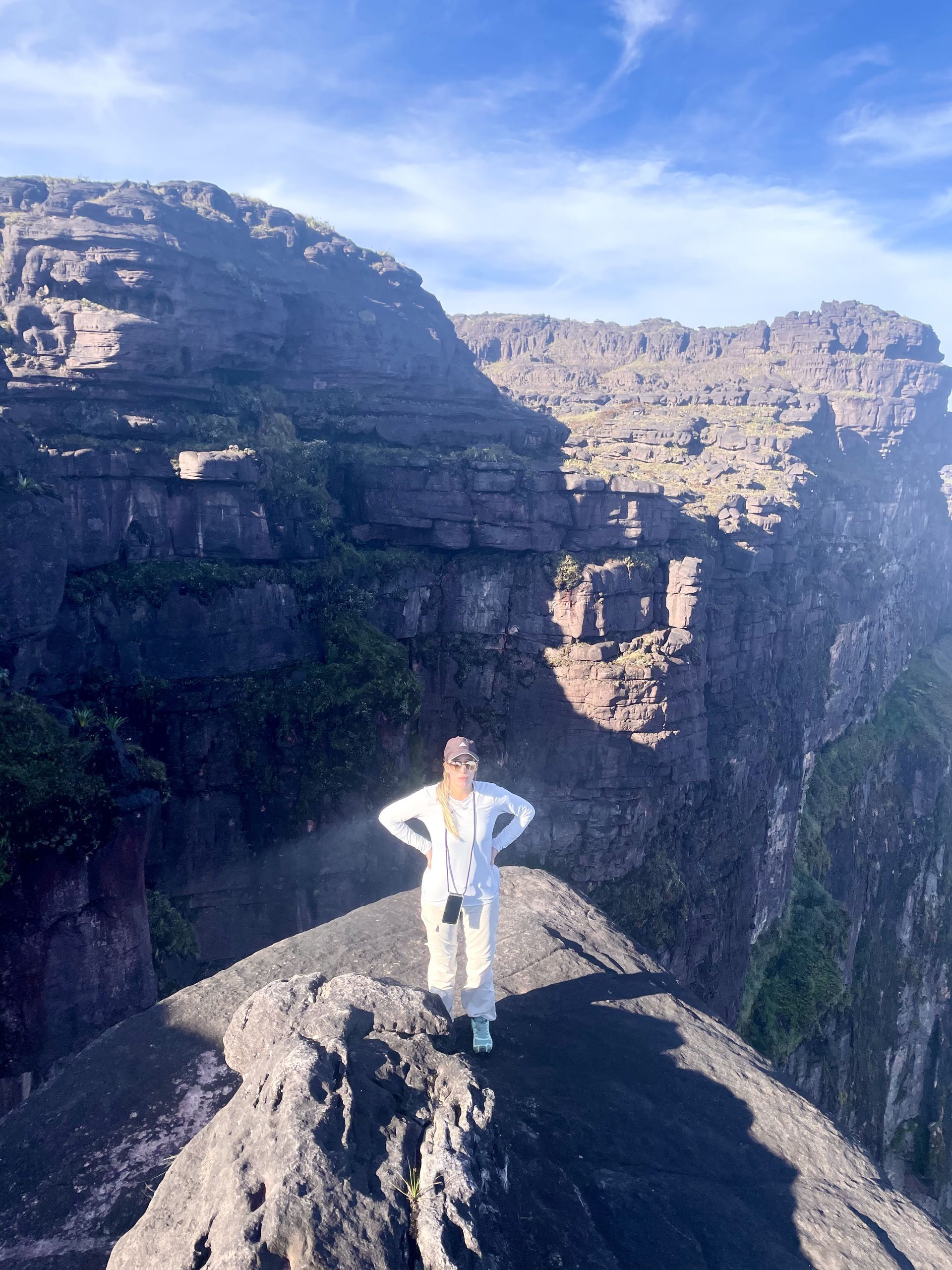 Woman stands on a rocky mountain ridge, arms akimbo, with steep cliffs and blue sky in the background.
