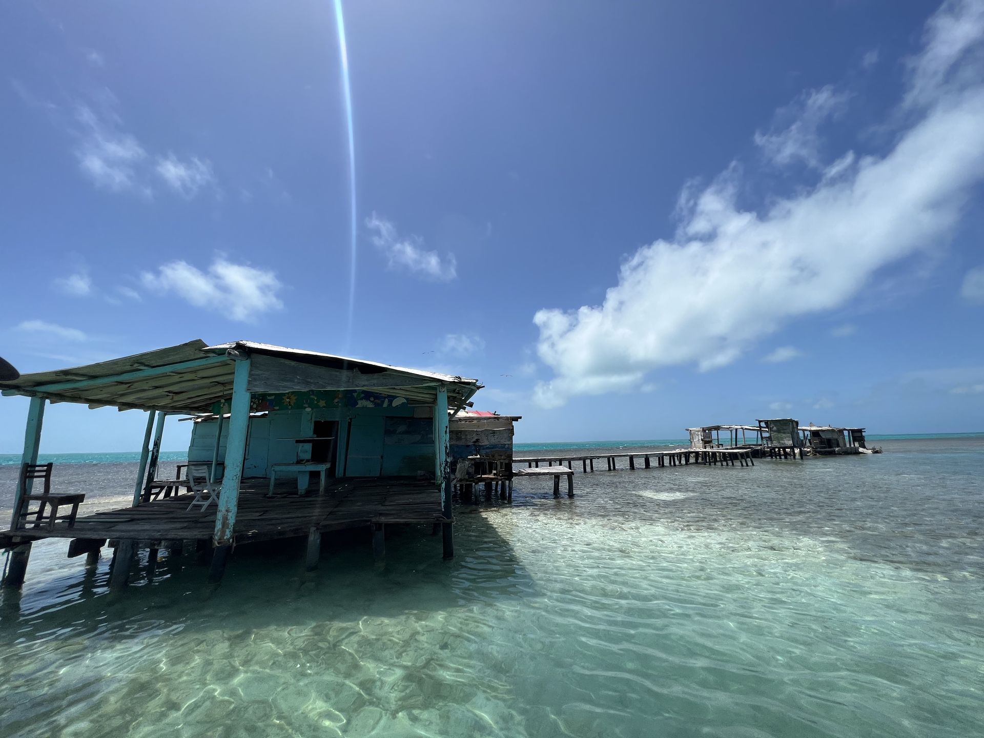 A weathered, light blue shack on stilts over shallow, clear water, with a pier leading to other structures under a bright blue sky.