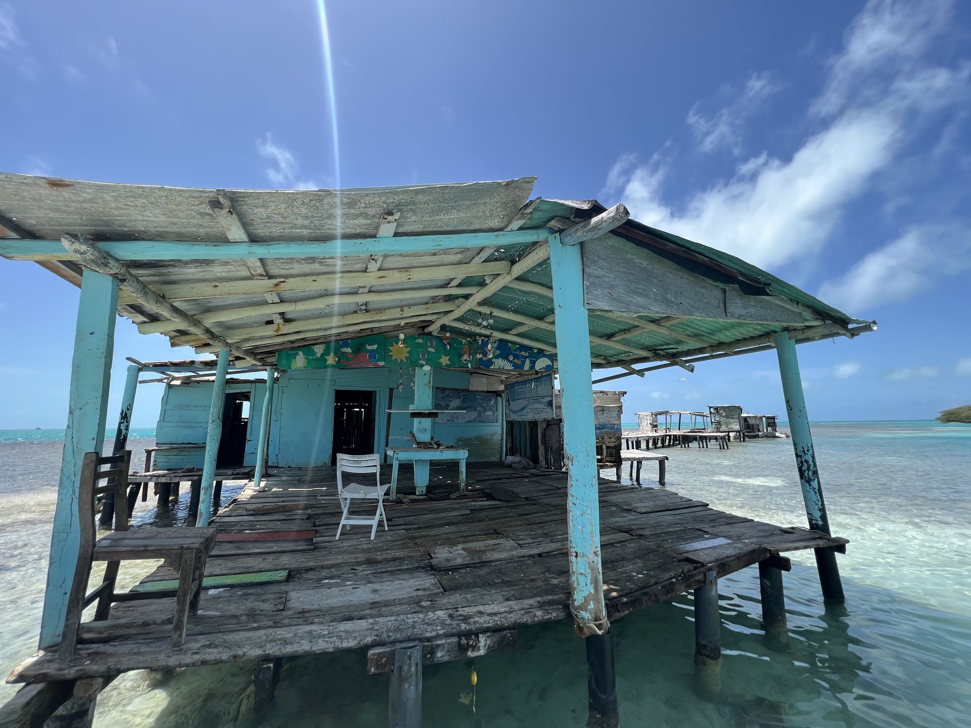 Dilapidated blue shack built over clear water with a blue sky backdrop. The shack features a partially collapsed roof and weathered wood.
