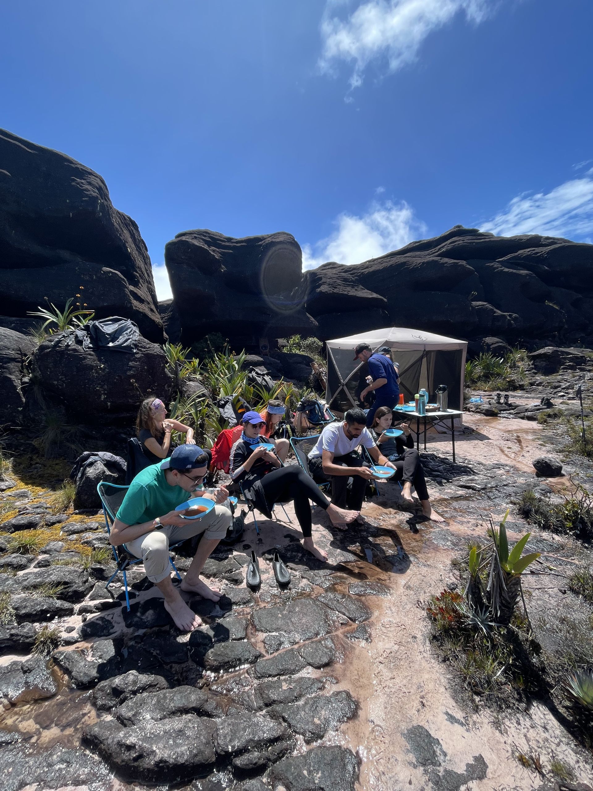 A group of people are resting and eating outdoors, possibly on a mountain. They are sitting on rocks near a small tent, under a bright blue sky.