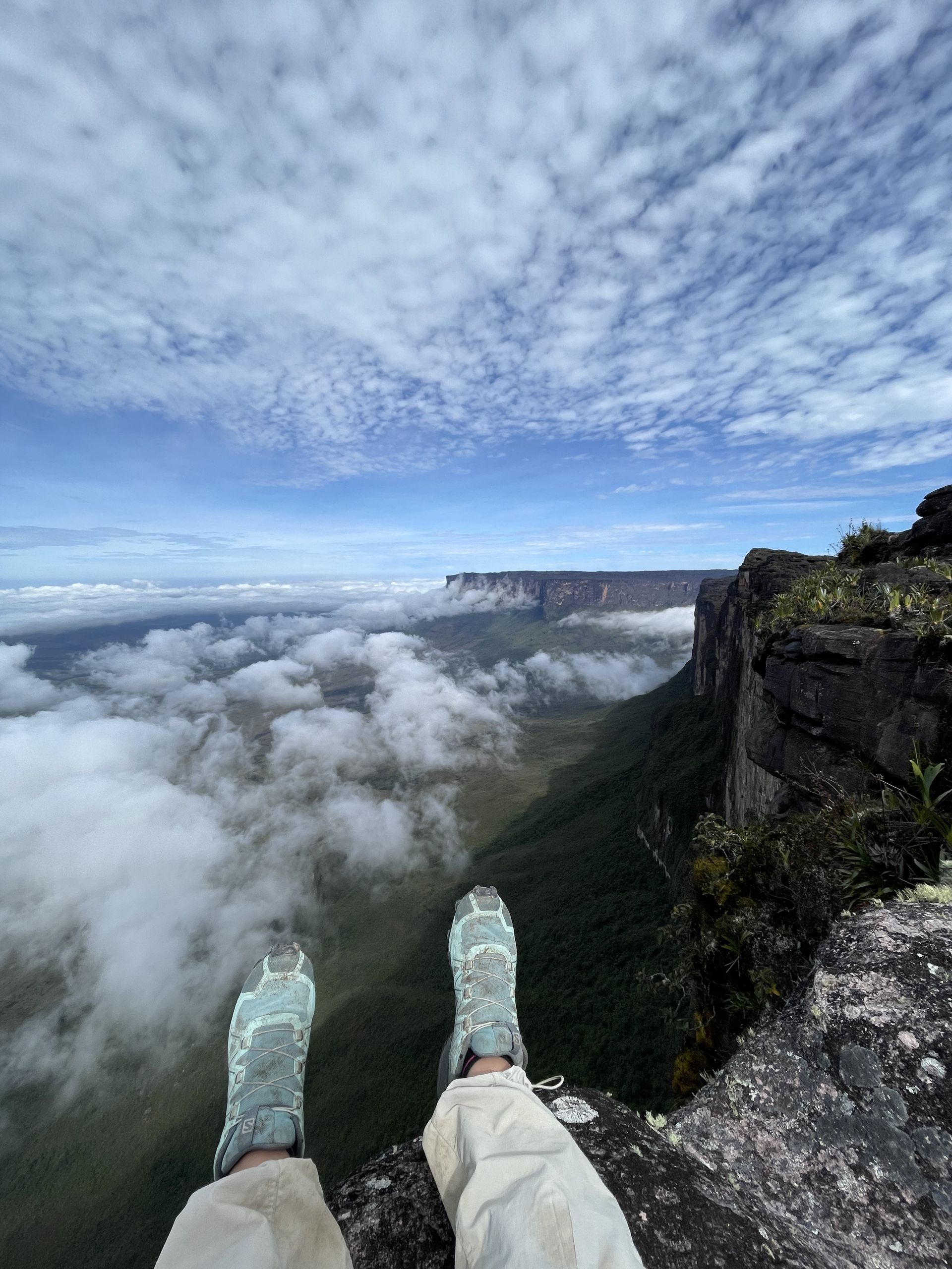Person sitting on a cliff overlooking a landscape of clouds, trees, and mesas. Sky is blue with scattered clouds; the person wears beige pants and light-colored hiking shoes.