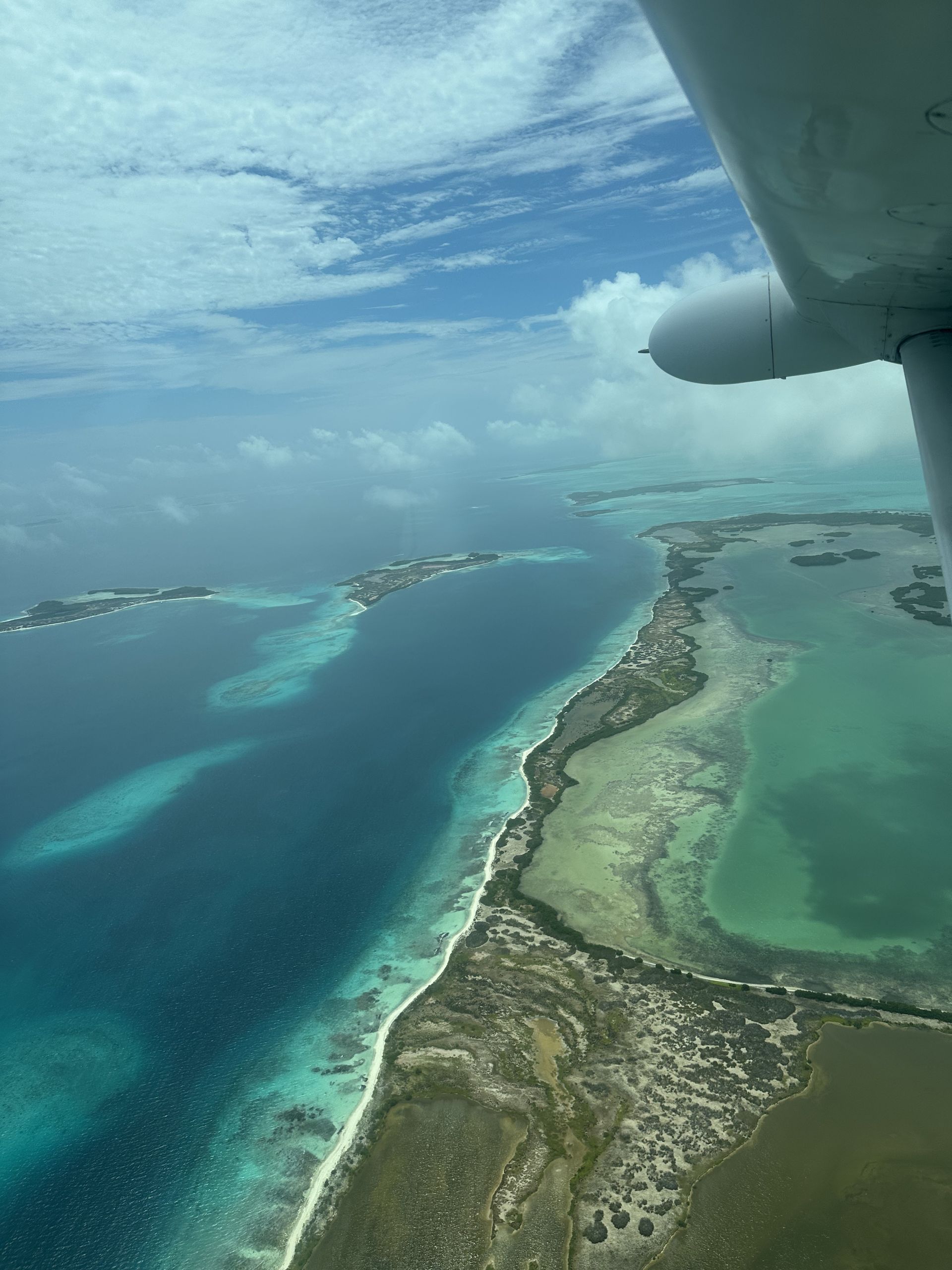 Aerial view of a turquoise ocean and land, possibly islands, under a partly cloudy sky. Taken from inside an airplane.