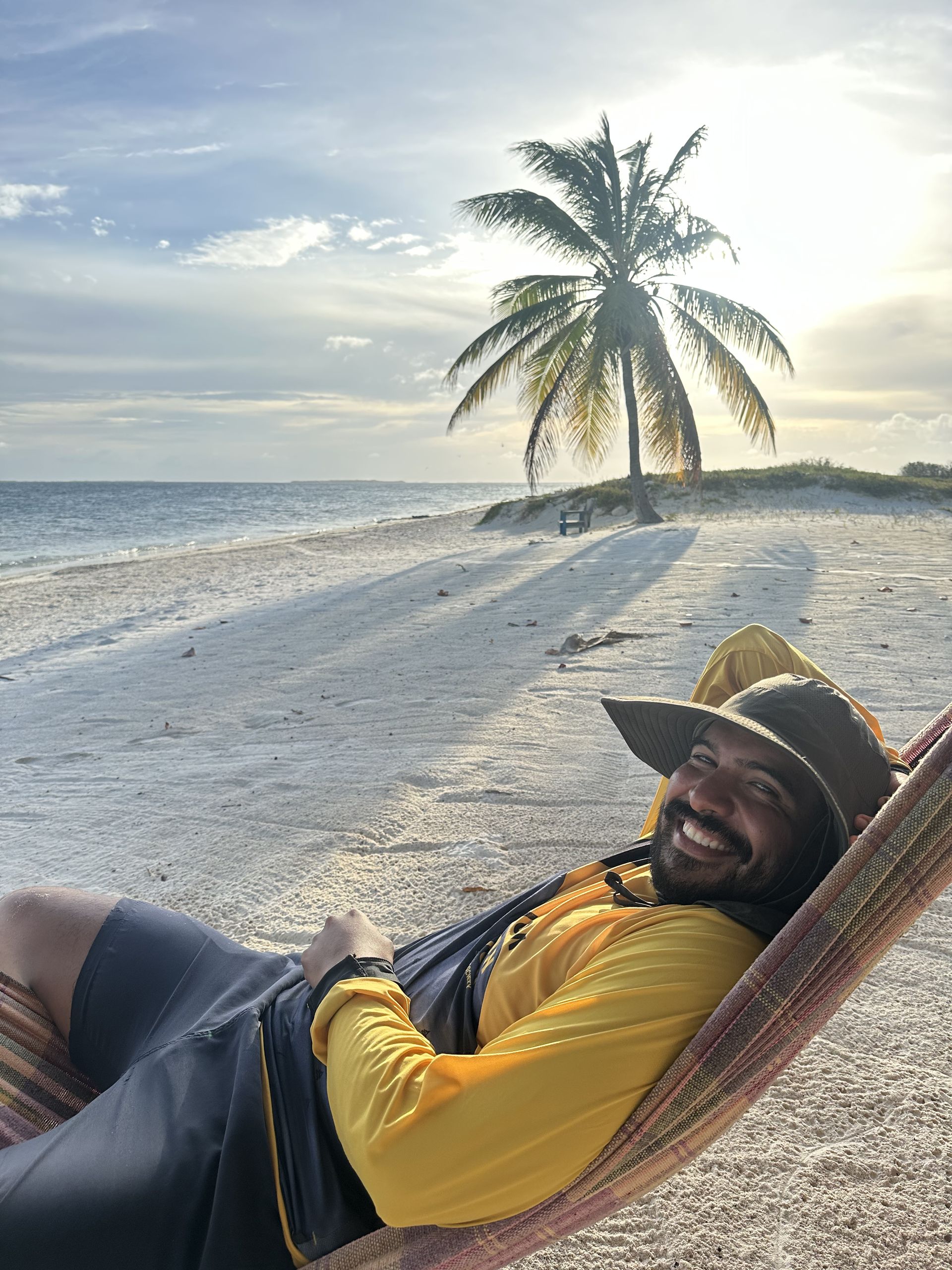 Person in a hammock smiles on a beach, palm tree in background, with a yellow shirt, hat, and shorts.