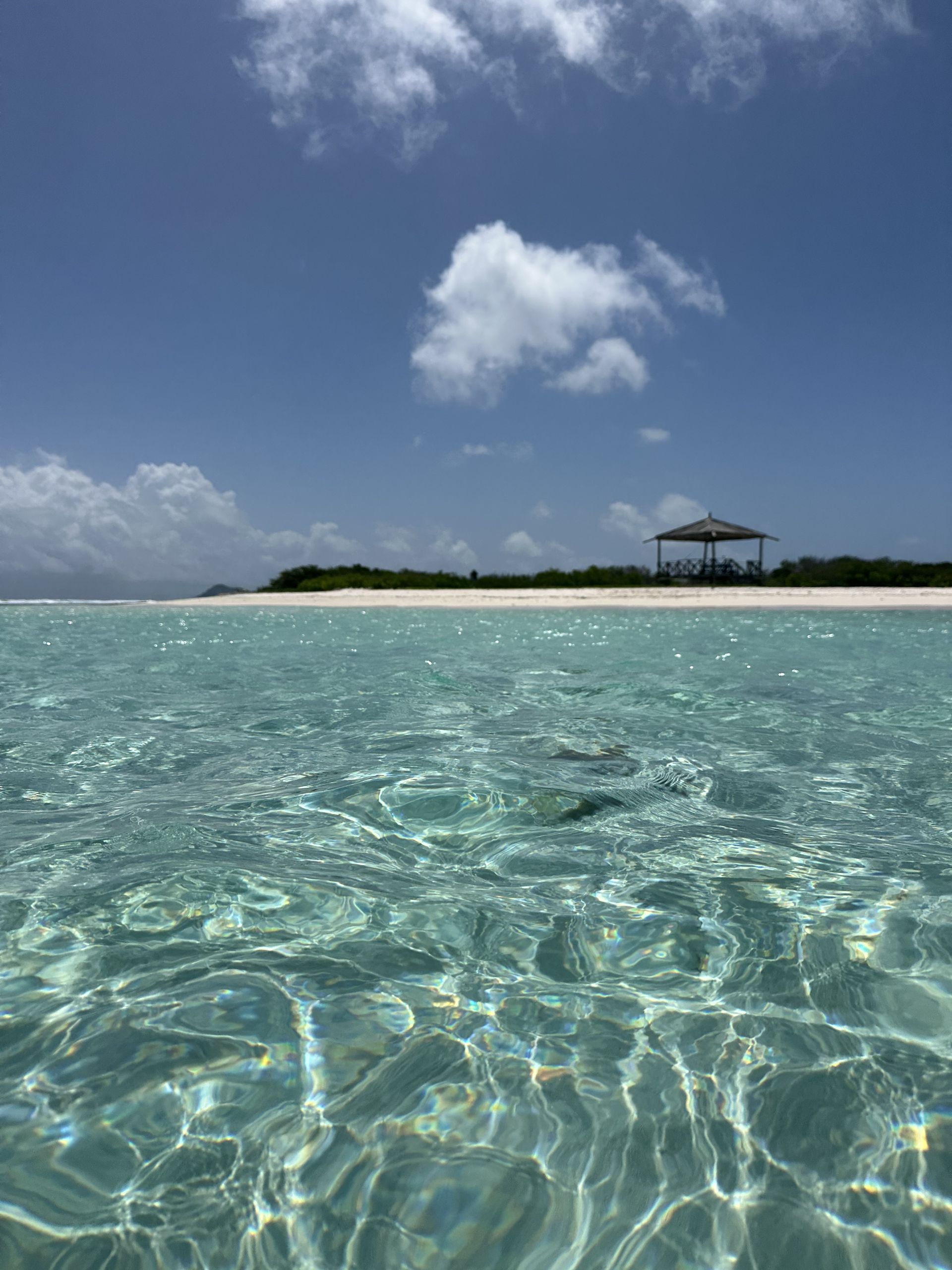 Clear turquoise water with sparkling sunlight, leads to a white sand beach and a gazebo on a sunny day.